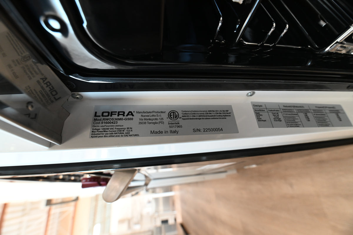A close-up of a Lofra product label sticker on the white inner frame of a dishwasher, with the black outer door visible above and a wooden floor in the background.