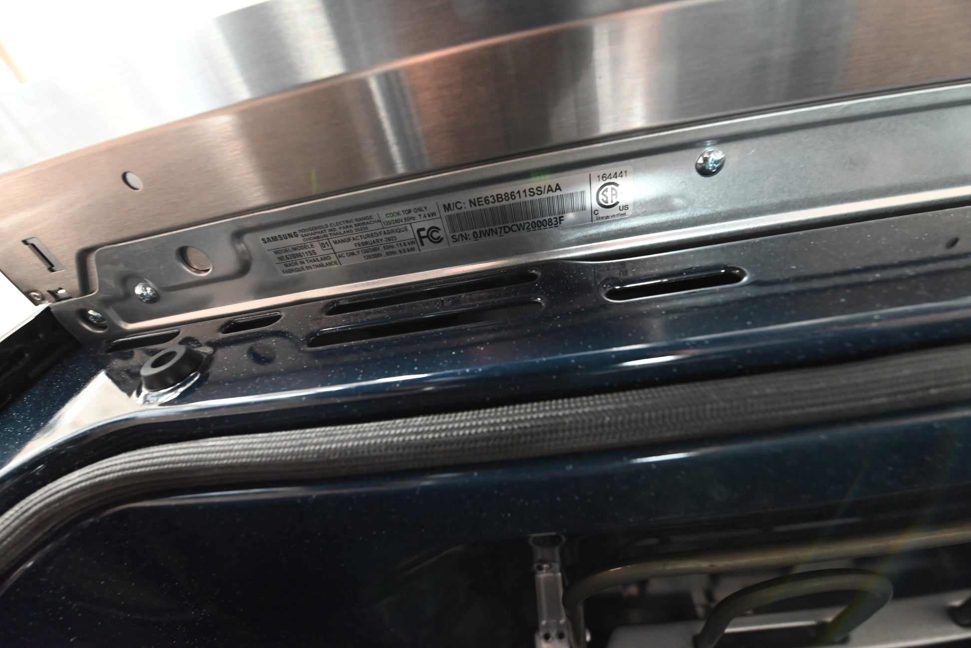 Close-up of the interior top edge of a dark appliance, showing a stainless steel panel, screws, a black rubber gasket, and a printed product label with regulatory text.