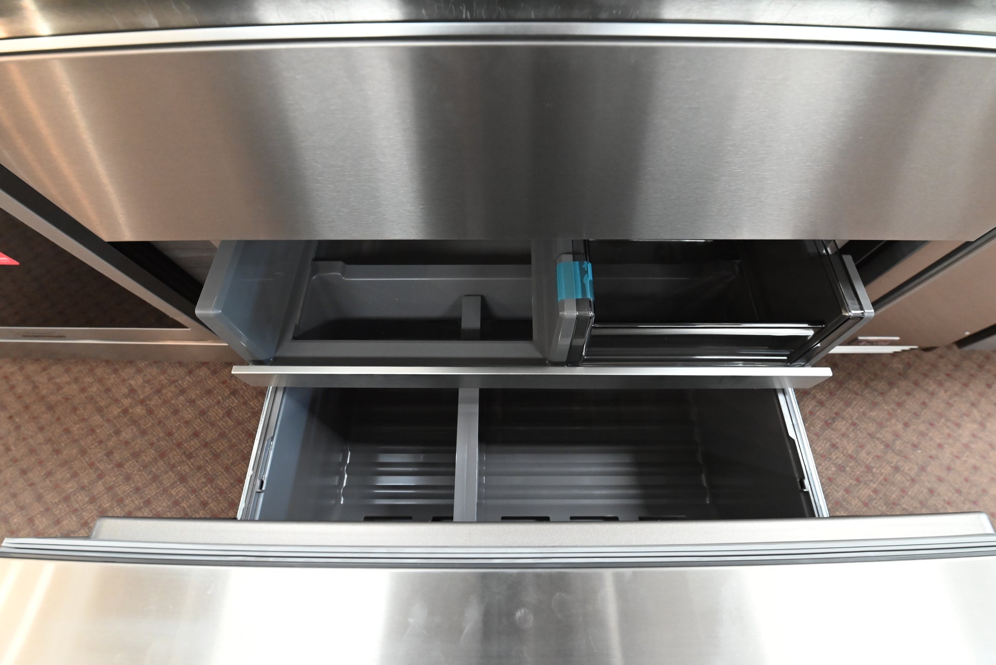 Close-up of a stainless steel refrigerator drawer system with one drawer pulled open, showing interior bins and a dark gray water filter cartridge with a blue tab.