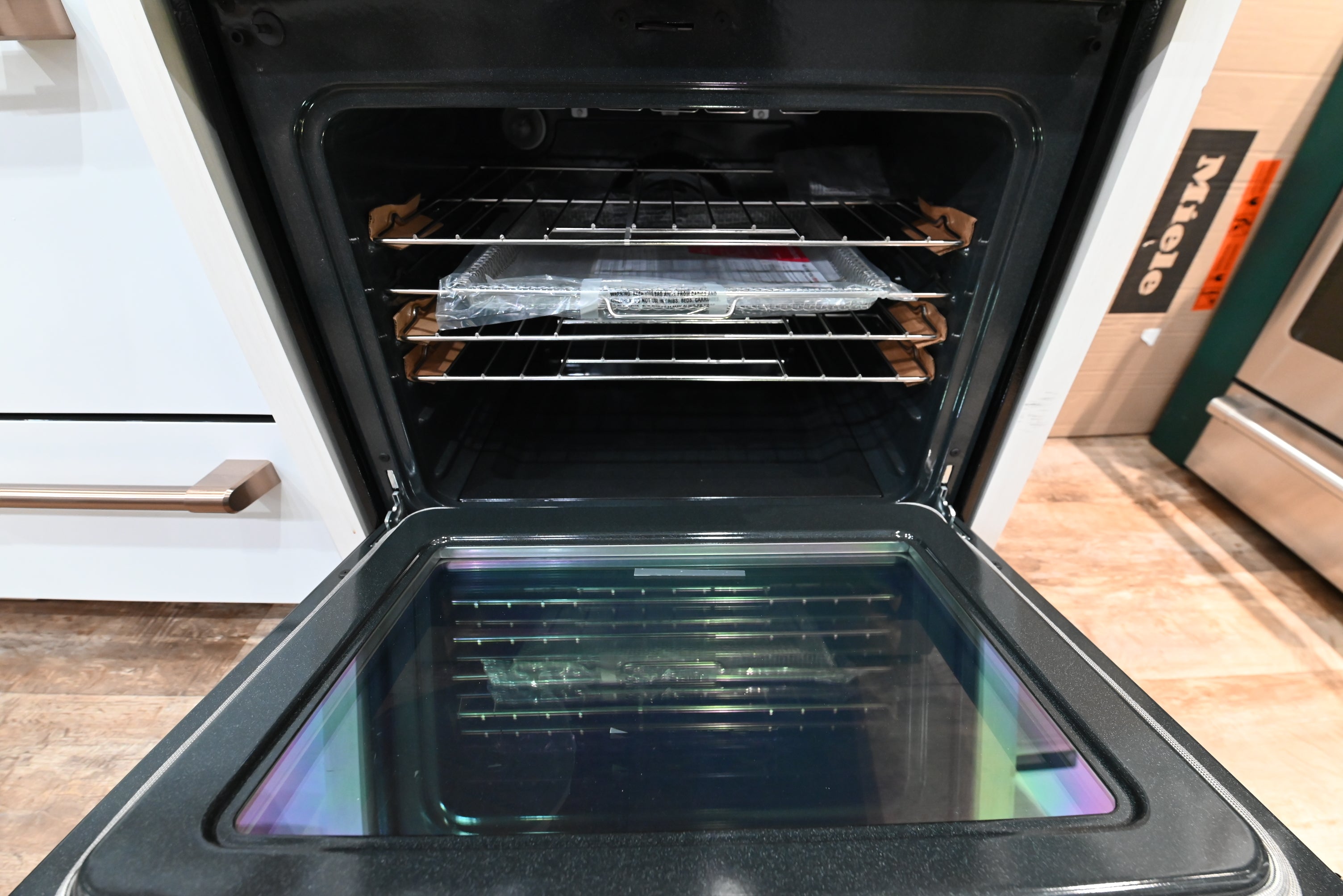 An open built-in oven with three racks inside, showing a plastic-wrapped item on one rack, set in a kitchen with white cabinetry and a wood floor.