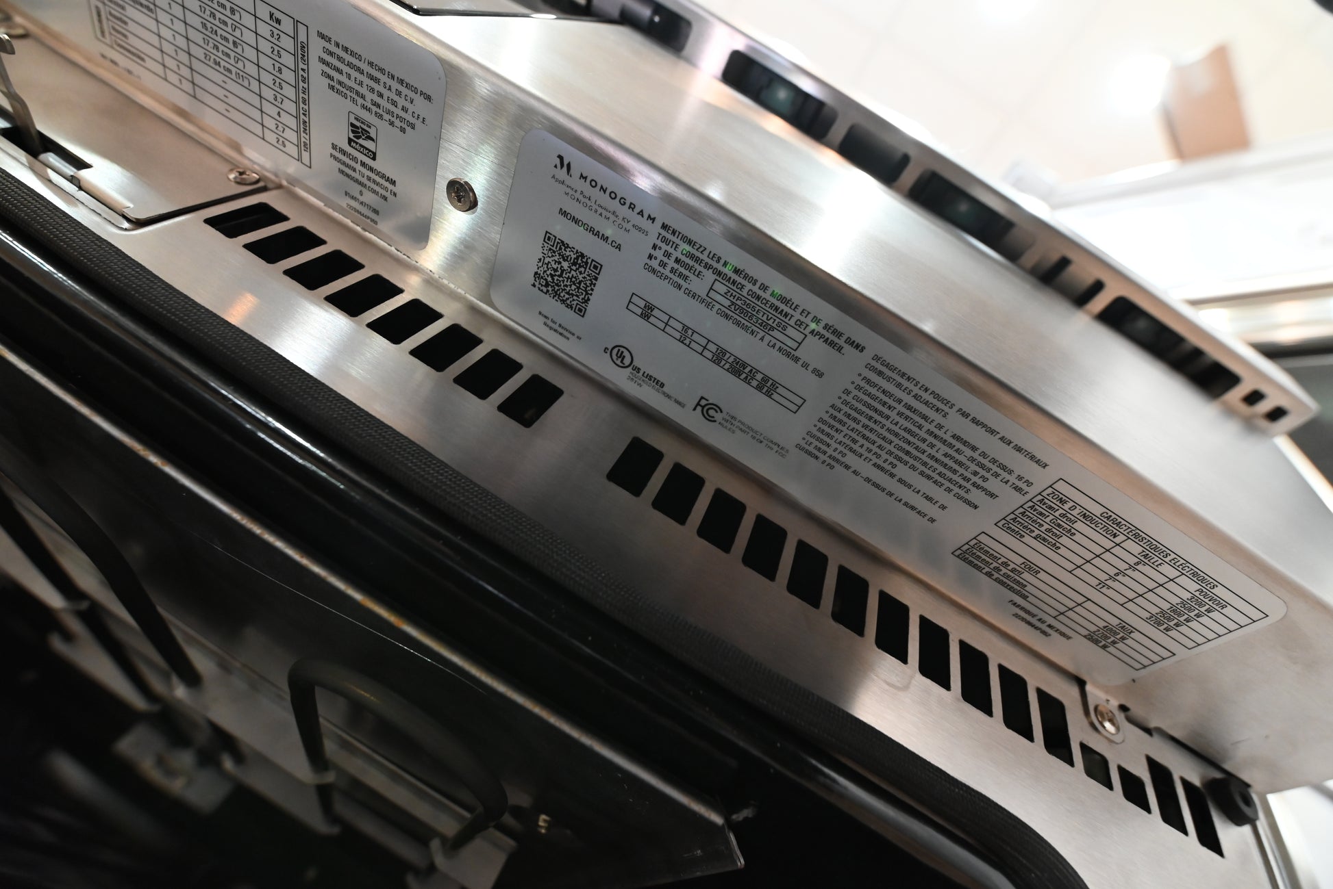 Close-up of the underside of a stainless steel appliance, showing a metal mounting bracket with printed specifications, a QR code, and ventilation slots.