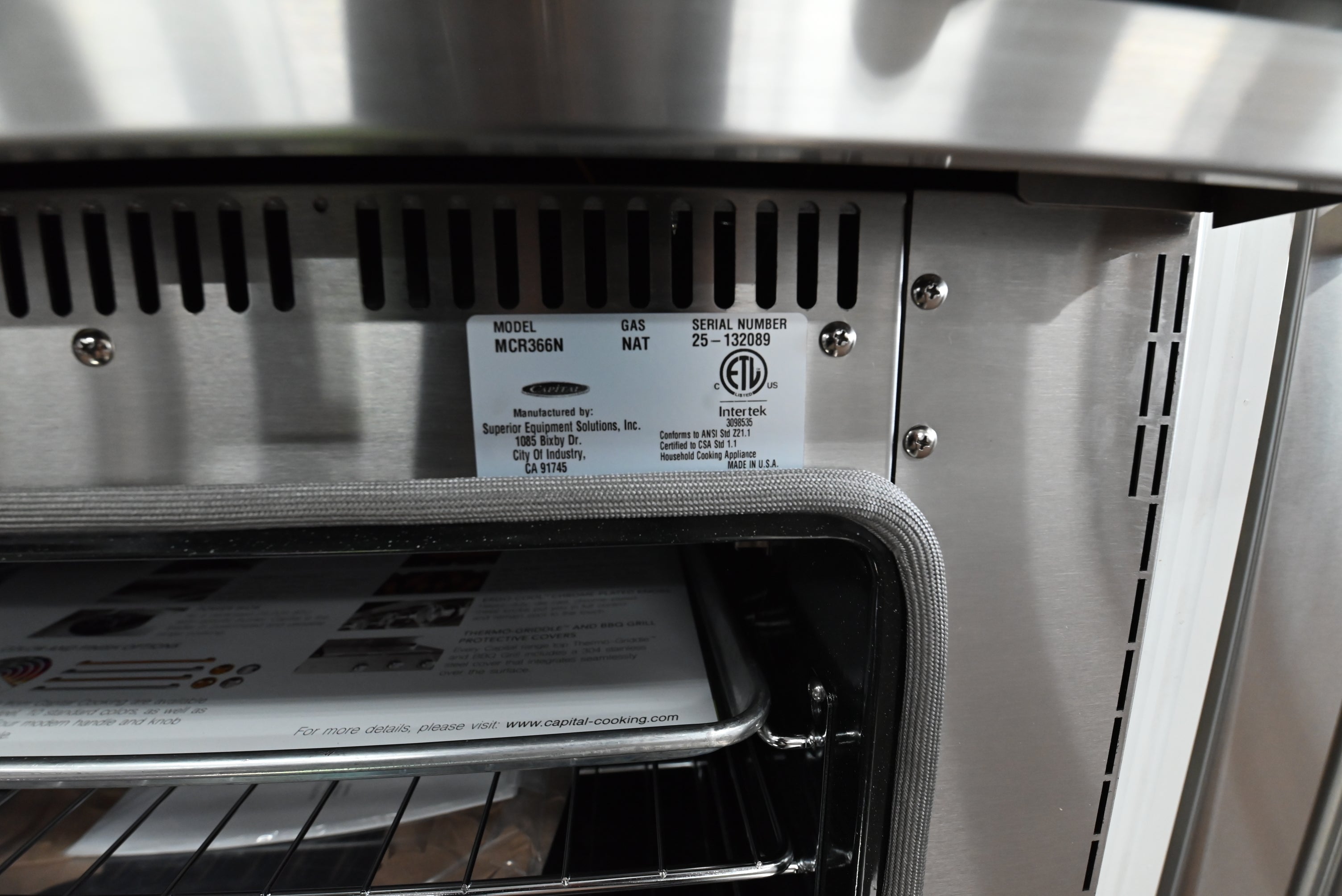 Close-up of the back panel of a stainless steel built-in oven, showing the model and maker label, vent slots, and the open door with interior racks.