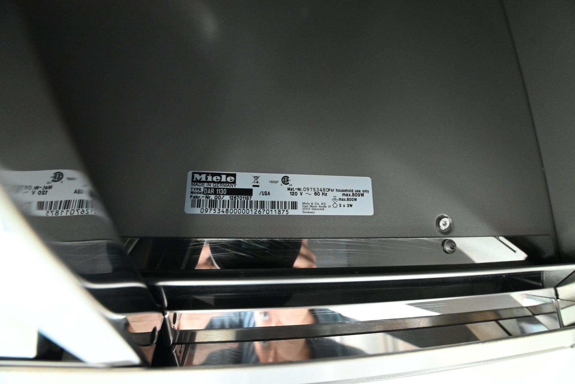 Close-up of the underside of a black appliance, showing a white Miele product label with barcode and model information, along with a reflective metallic handle.