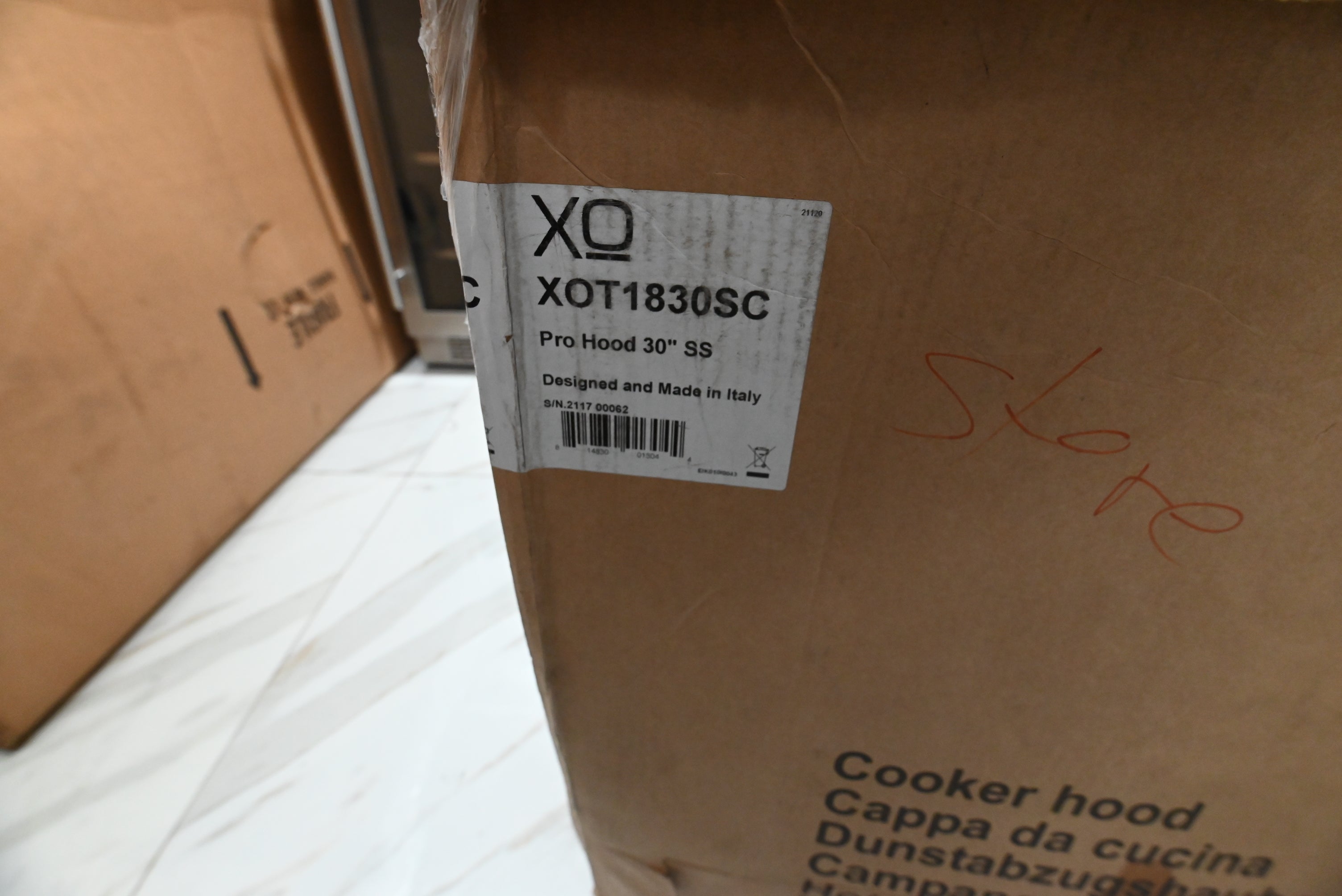 A close-up of a brown cardboard shipping box with a white label reading XO XOT1830SC Pro Hood 30" SS, photographed in a warehouse or storage area.