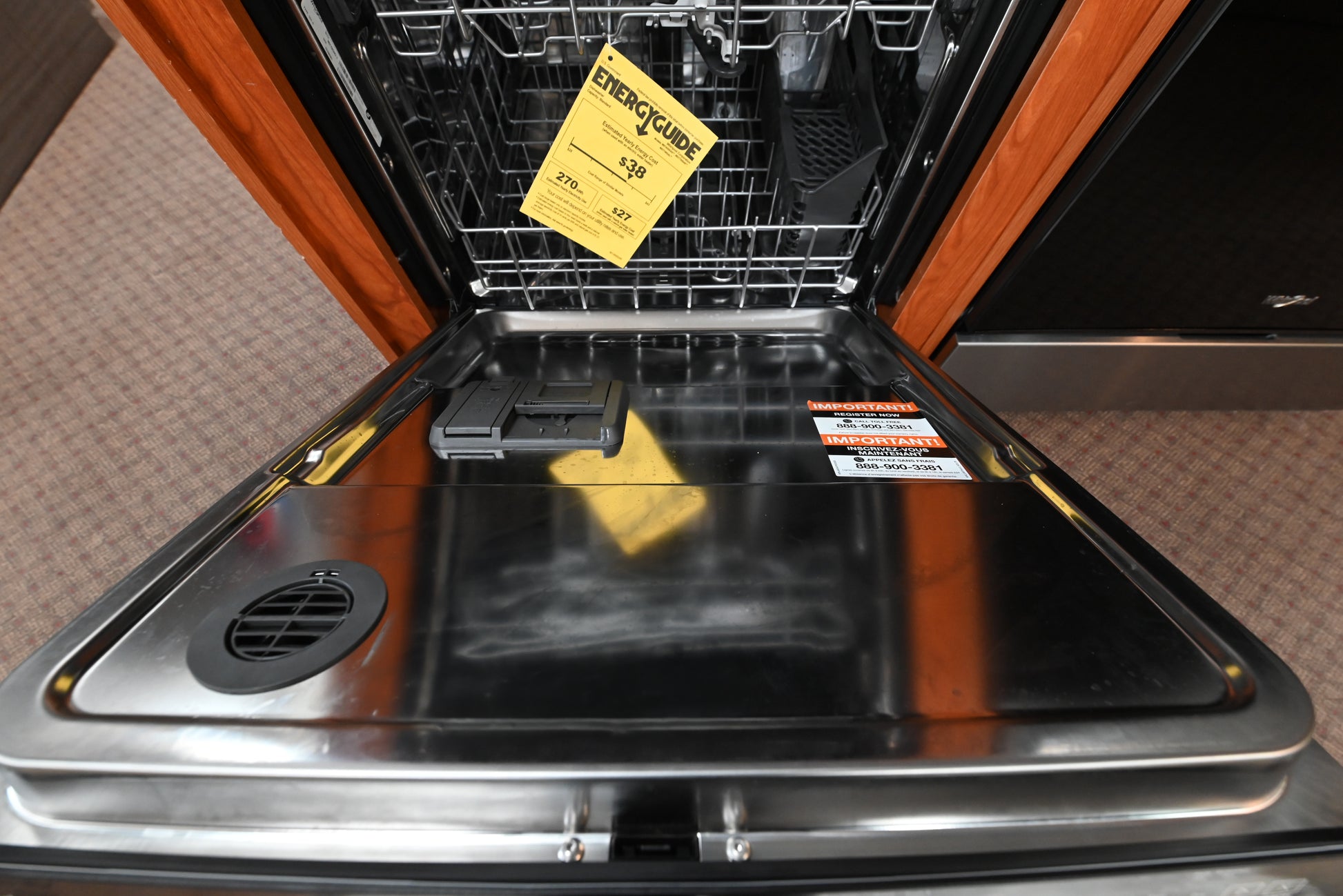An open stainless steel built-in dishwasher with a yellow EnergyStar price tag hanging in the upper rack, shown between wood cabinet panels.