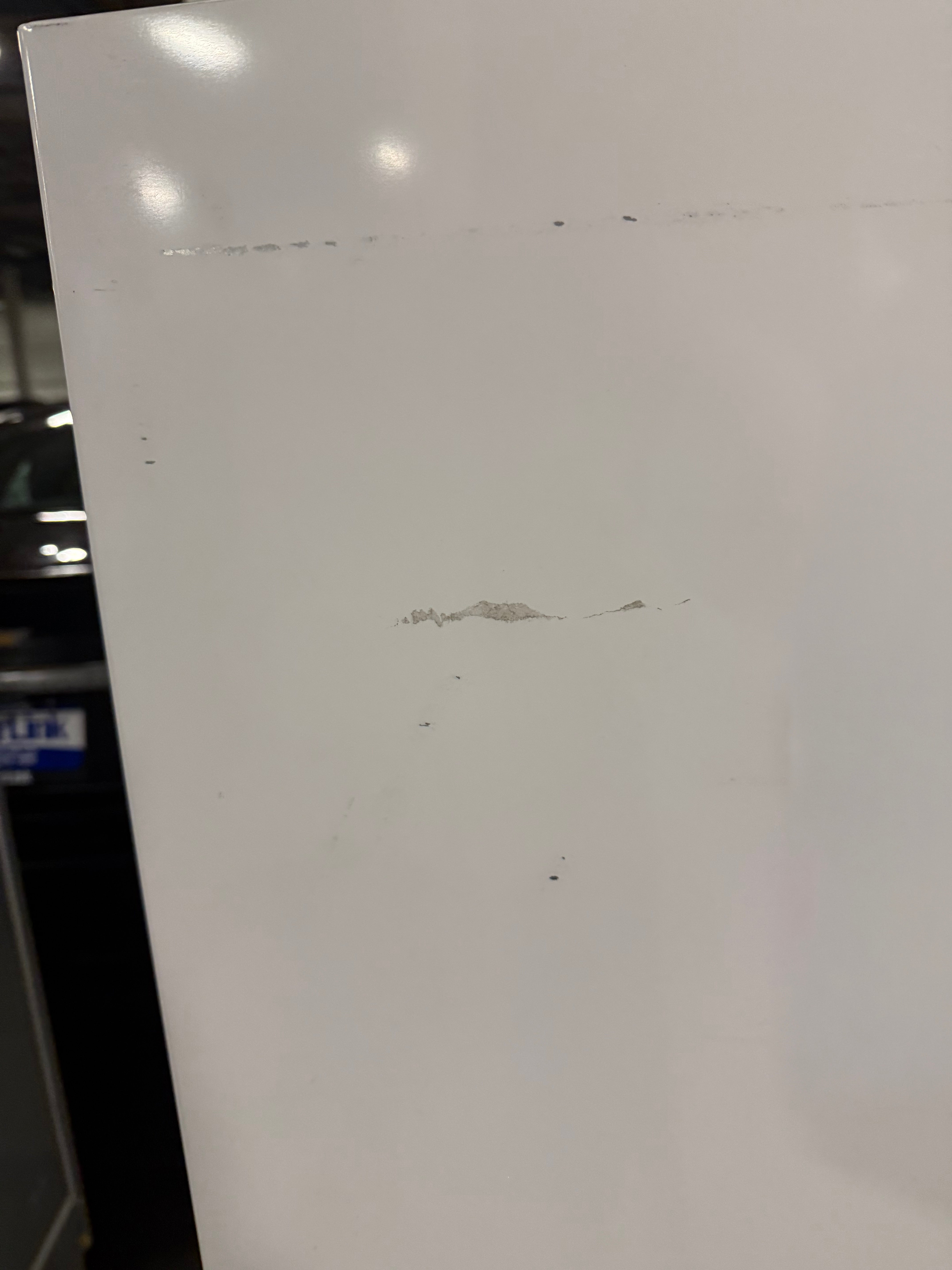 A close-up of a white panel or board with a few small scratches and marks, photographed indoors with some reflections and a dark background visible on the left.