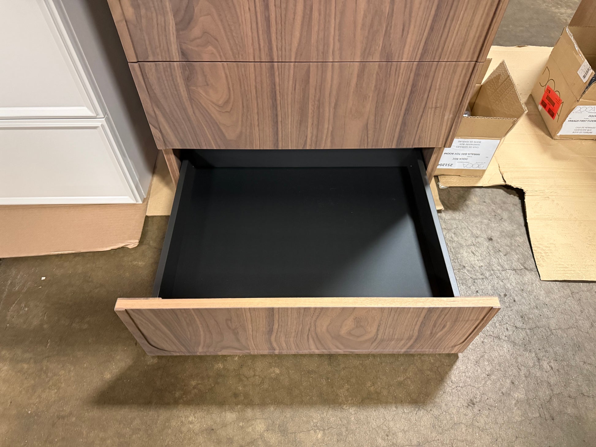 A close-up of a wooden drawer with the bottom pull-out drawer open, showing a black interior. The drawer has a medium brown wood grain finish and is set against a warehouse-like background with concrete floors and cardboard boxes.