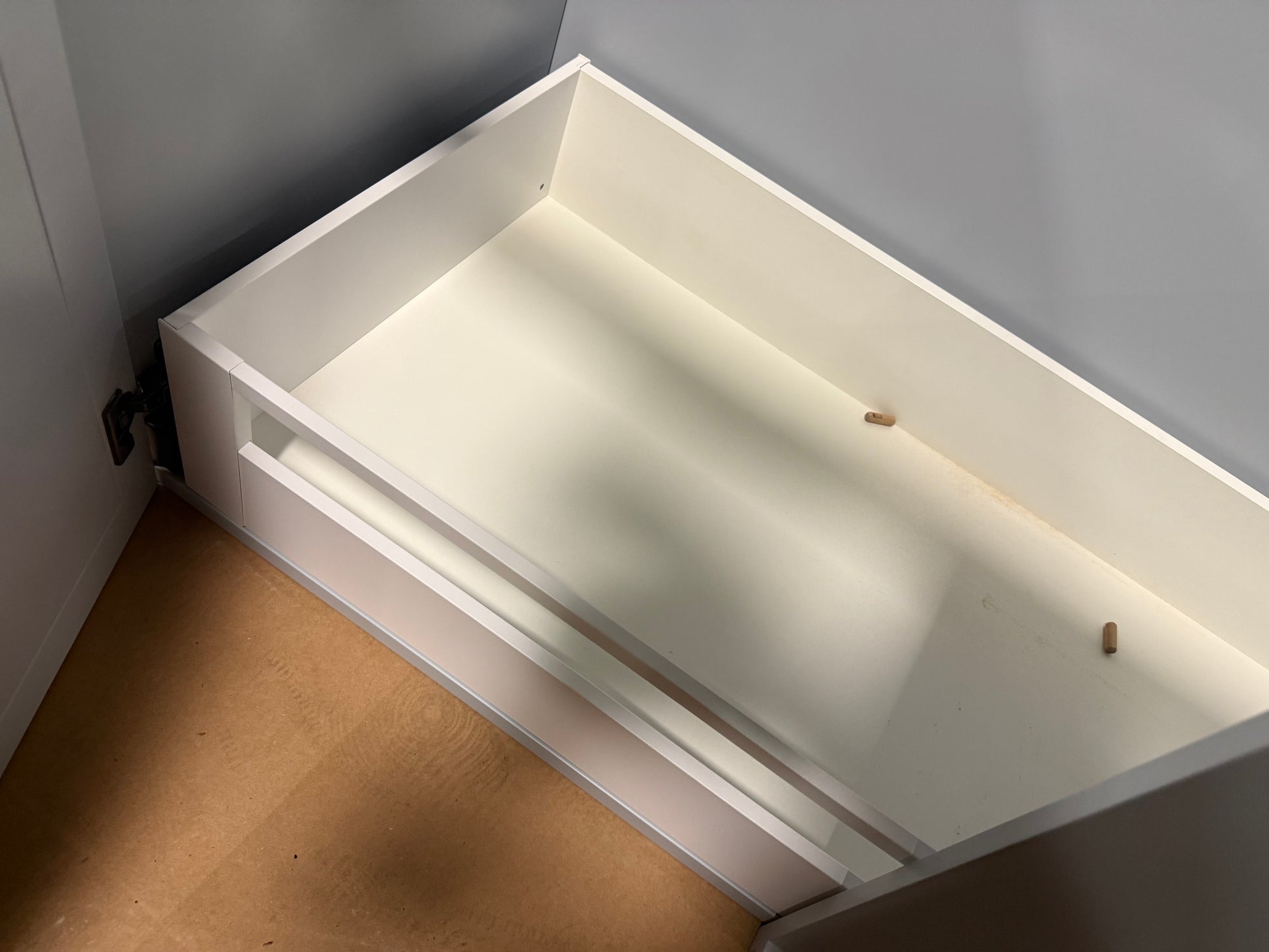 A white drawer or storage compartment shown from an angled top-down view, set against a gray wall and light wood floor.