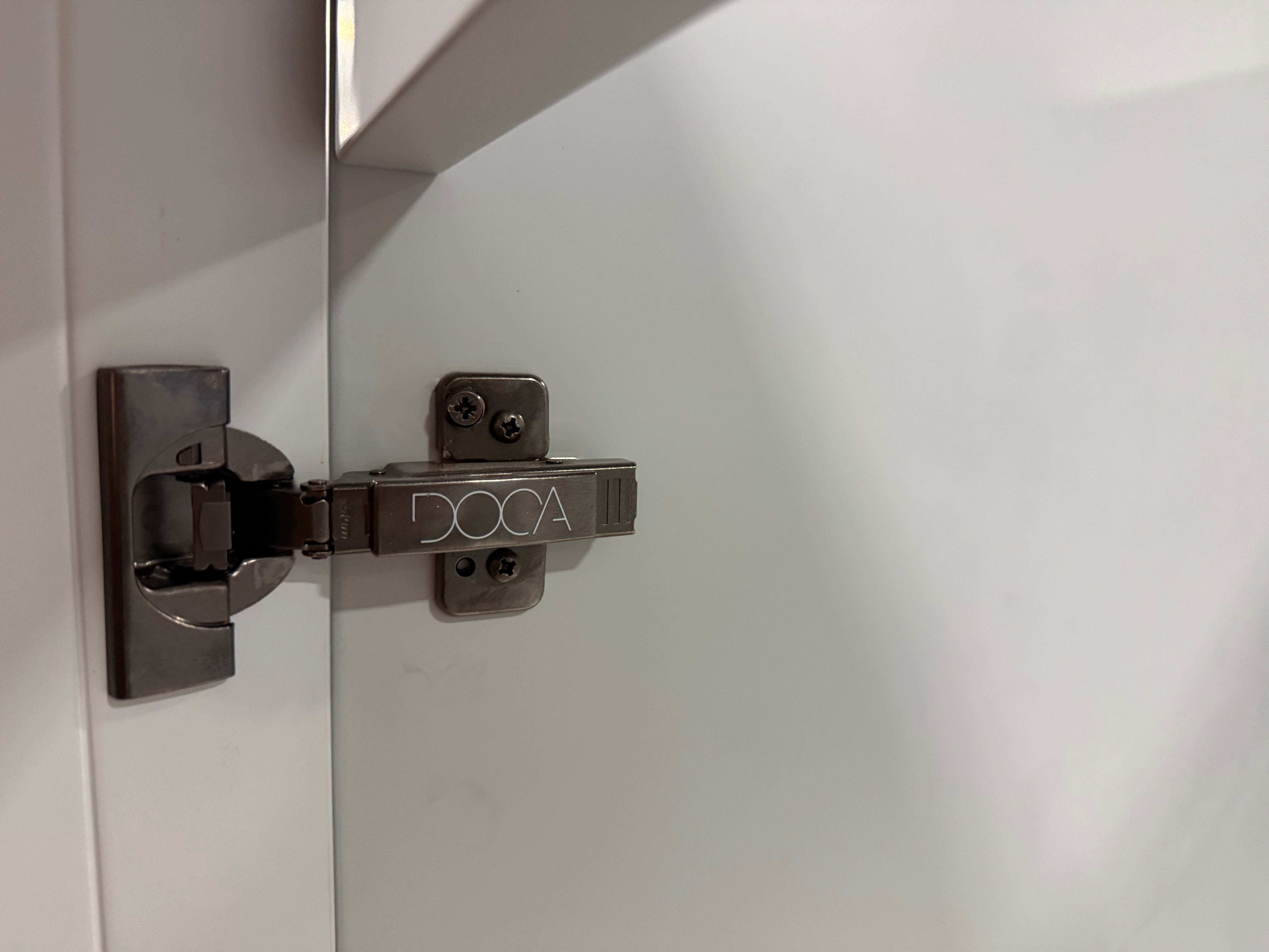 A close-up of a DOCA brand metal cabinet hinge mounted on a white cabinet door next to a shelf.
