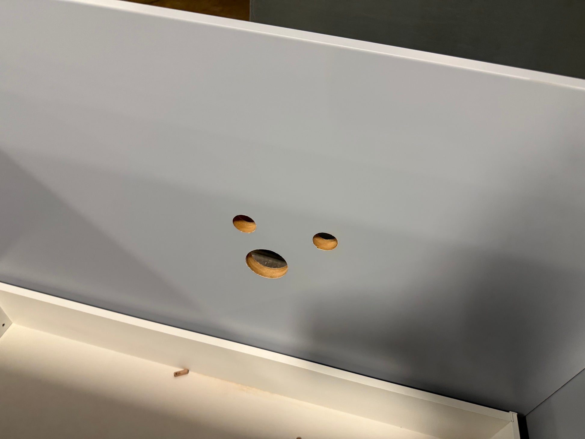 Close-up of a white wooden furniture piece, likely a cabinet or shelf, showing three circular screw holes on the front panel and the underside of the frame.