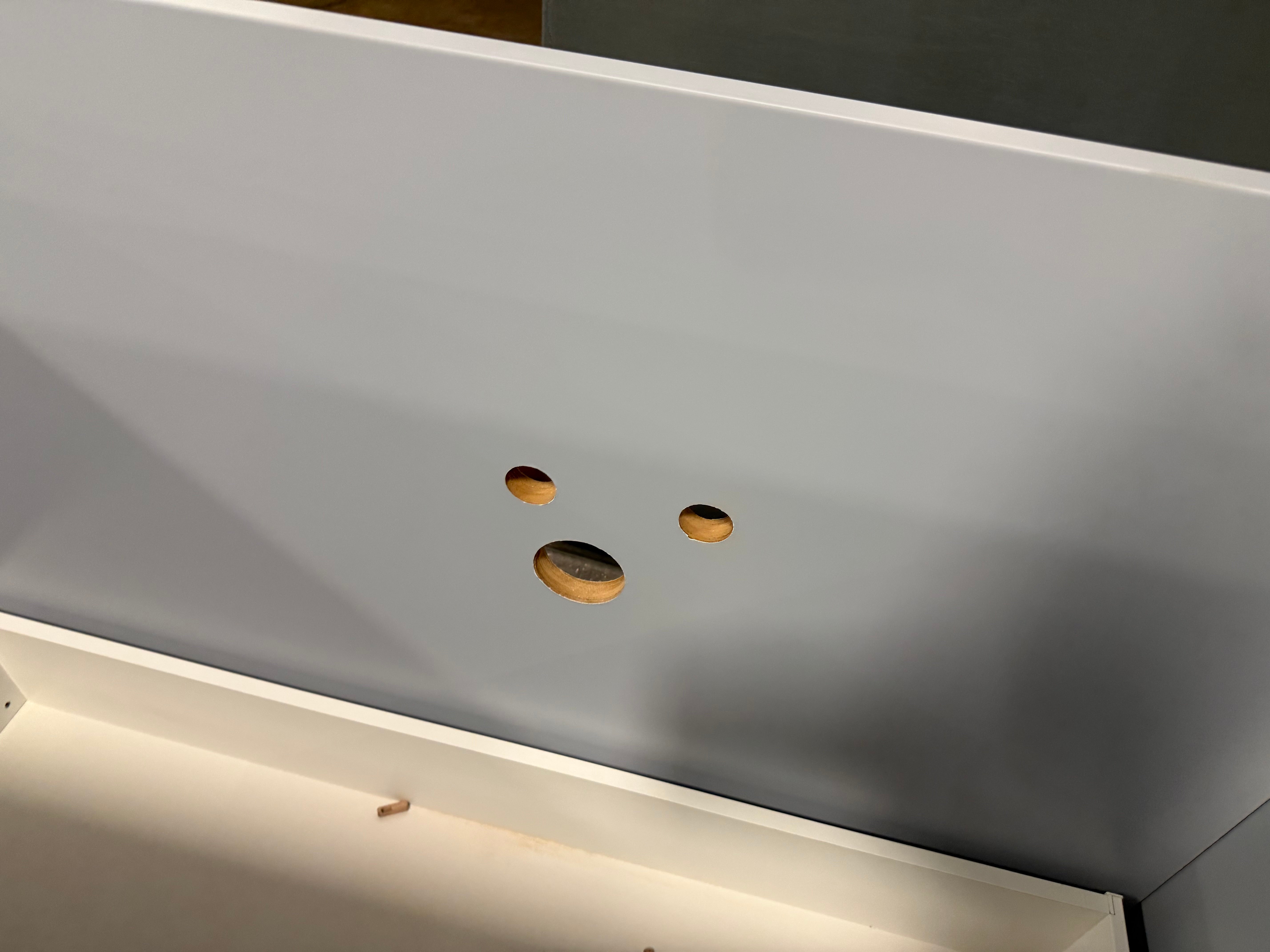 Close-up of a white wooden furniture piece, likely a cabinet or shelf, showing three circular screw holes on the front panel and the underside of the frame.