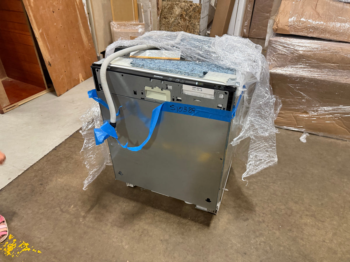 A stainless steel appliance, likely a dishwasher or similar household machine, wrapped in clear bubble wrap and secured with blue tape, sitting on a concrete floor in a warehouse-like setting.