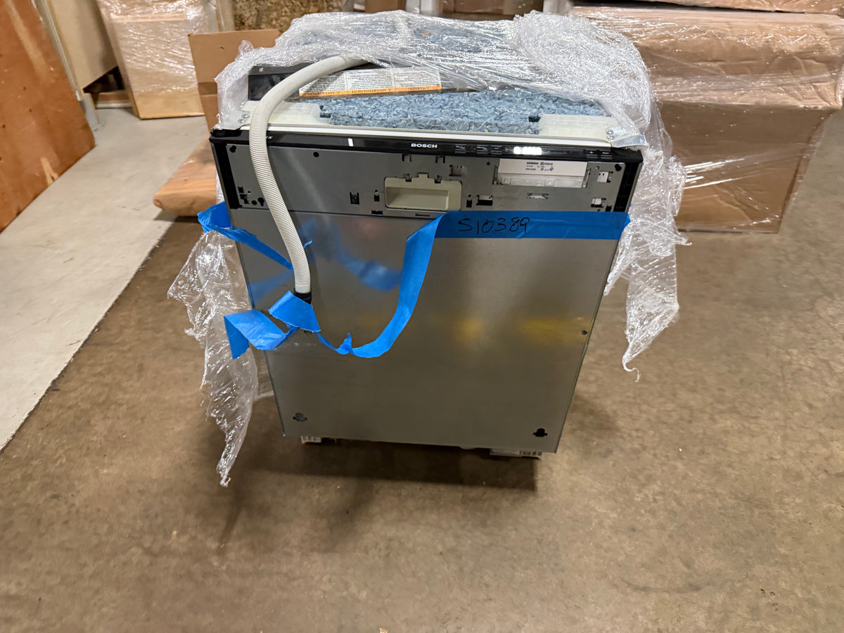 A stainless steel built-in dishwasher, shown from the back, wrapped in clear bubble wrap with blue tape and a white water supply line. The appliance is photographed on a concrete floor in what appears to be a warehouse or storage area.