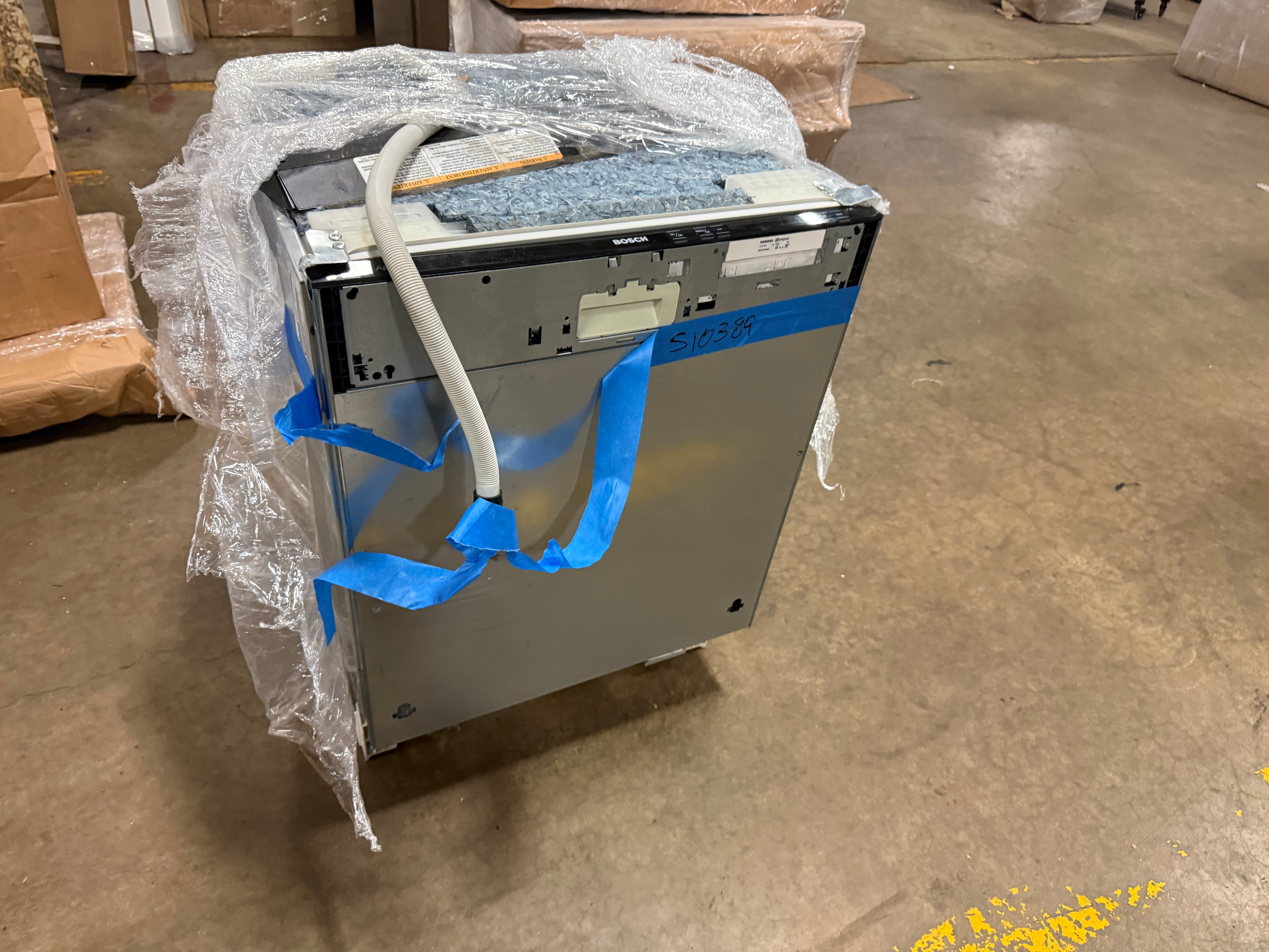 A stainless steel dishwasher, likely a Samsung model as indicated by the visible 'SAMSUNG' label, wrapped in clear bubble wrap and secured with blue tape on a concrete warehouse floor.
