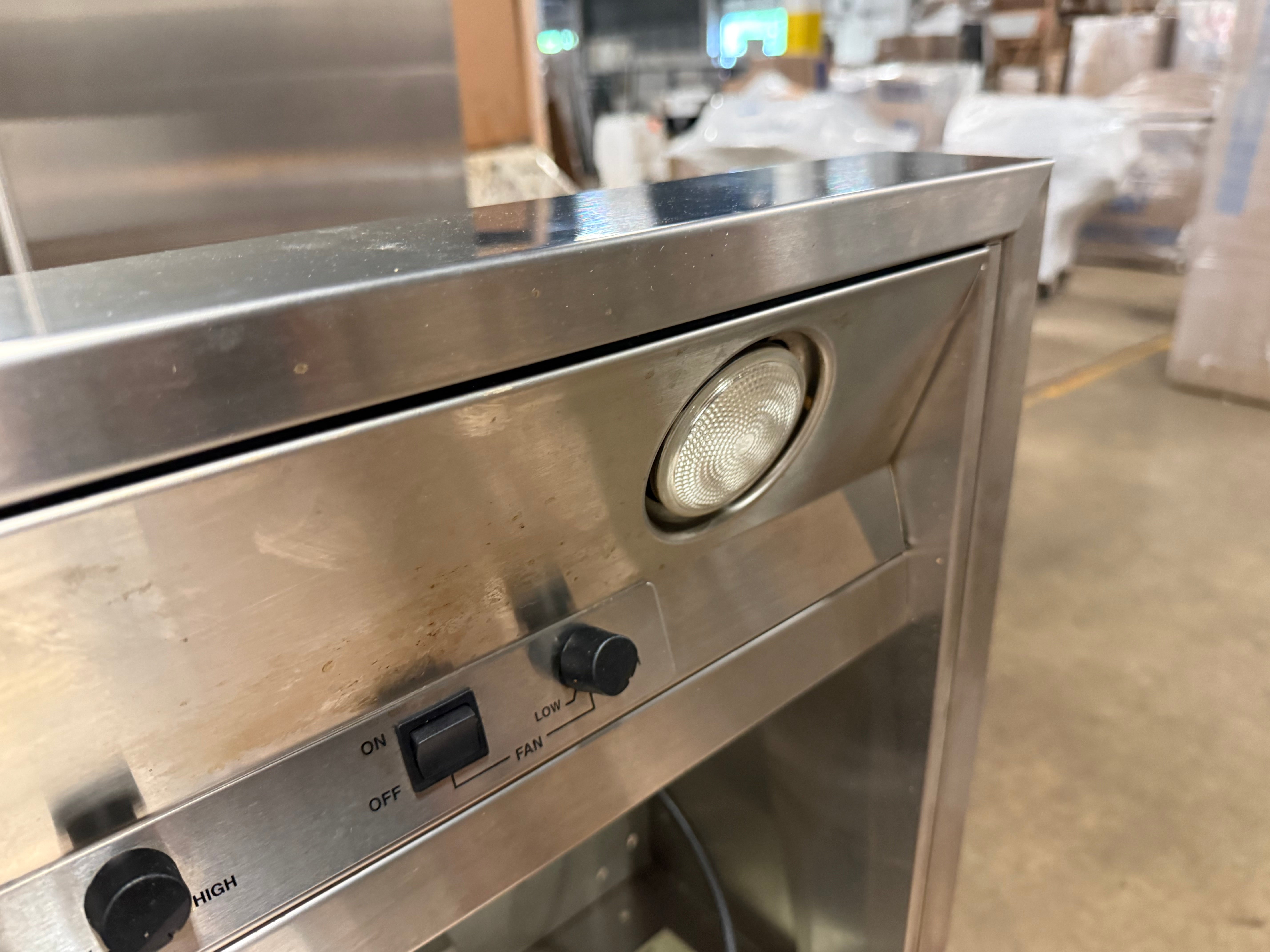 Close-up of a stainless steel commercial appliance control panel with a round light, switch, and dial, photographed in a warehouse setting.