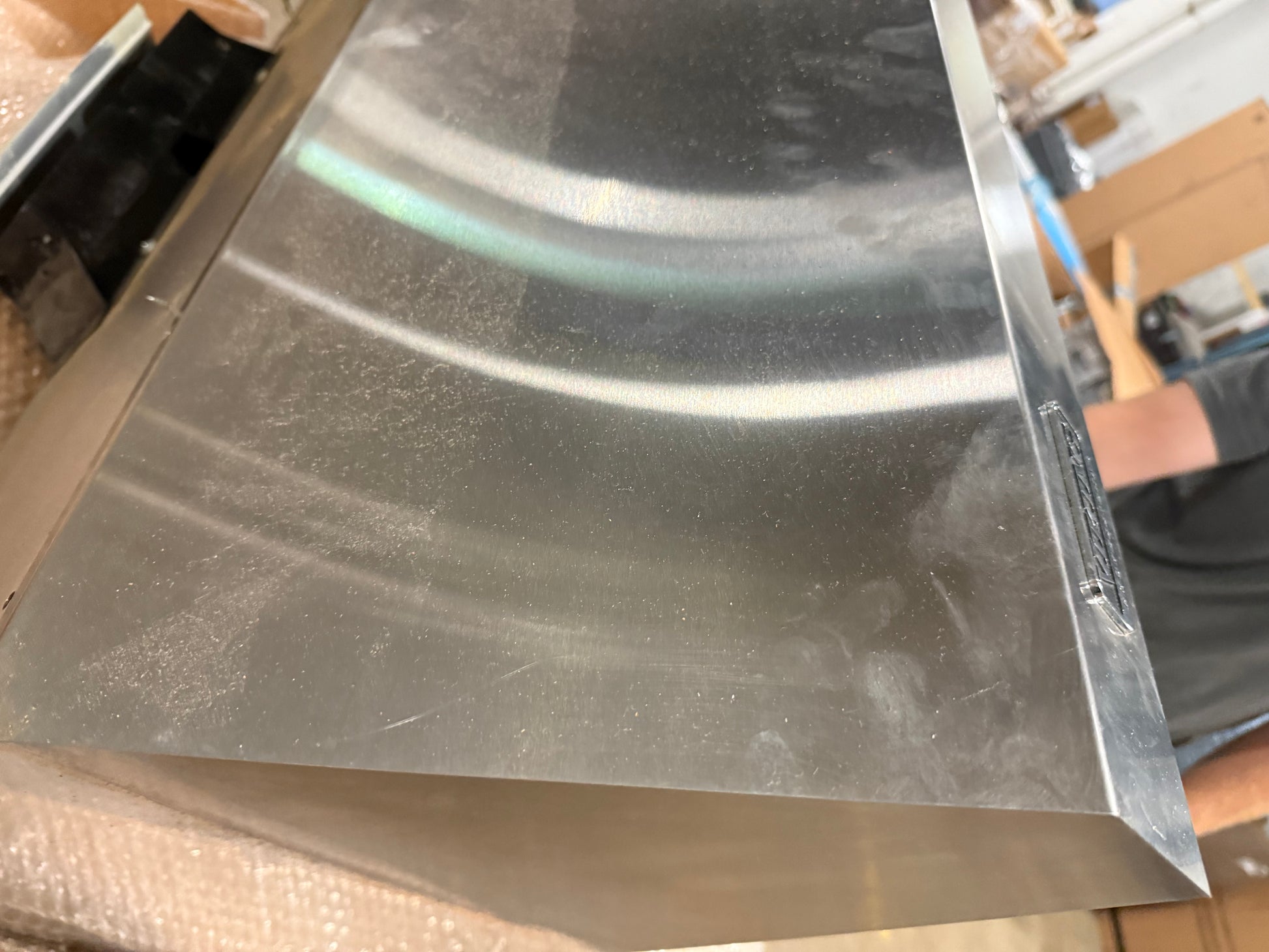 A close-up of a brushed stainless steel panel, likely an appliance component or cabinet front, showing a reflective metallic surface with dust and scratches.