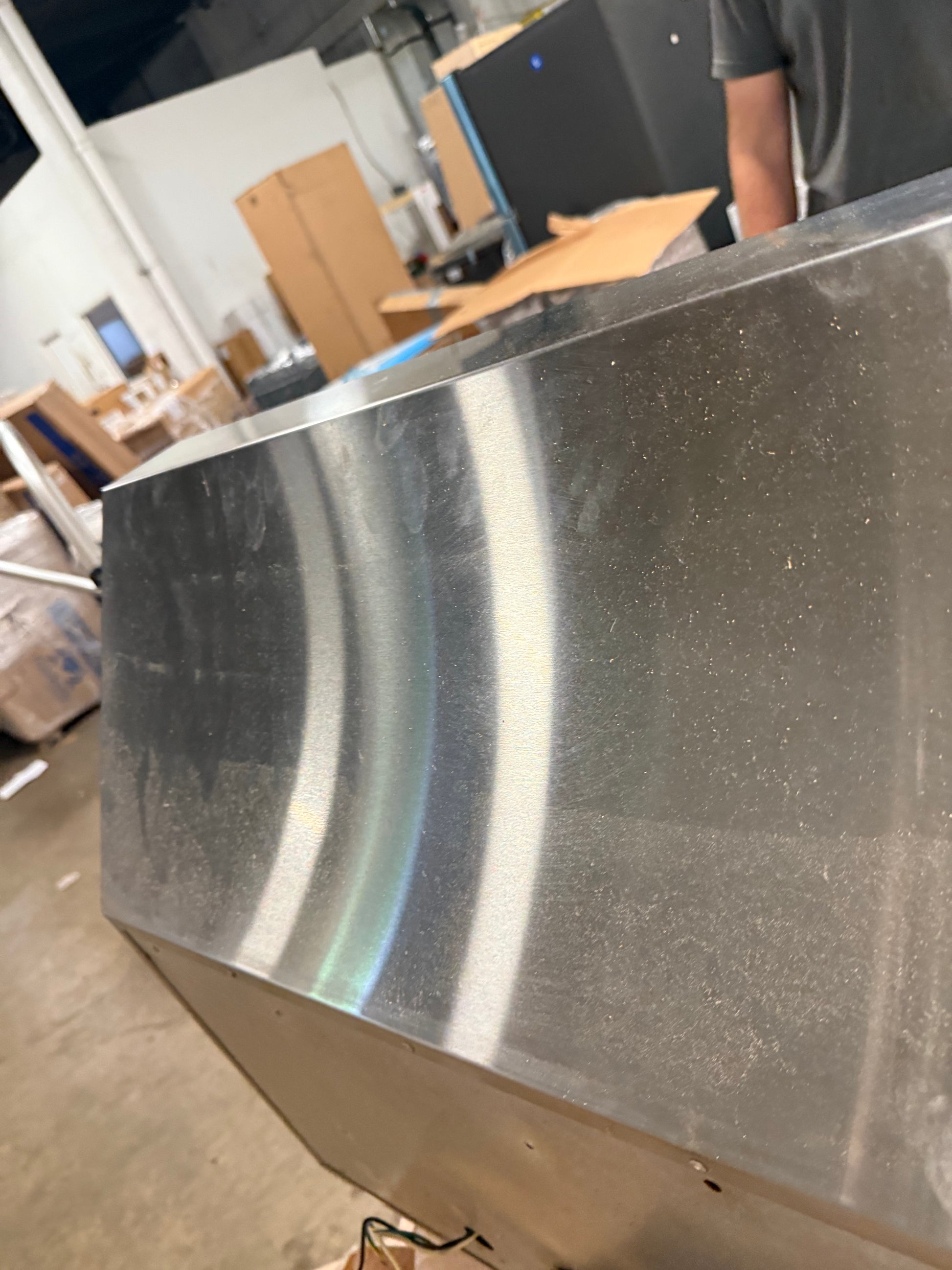 A close-up of a large, brushed stainless steel metal panel with a curved reflective surface, set in a workshop or warehouse environment with cardboard boxes and a person partially visible in the background.