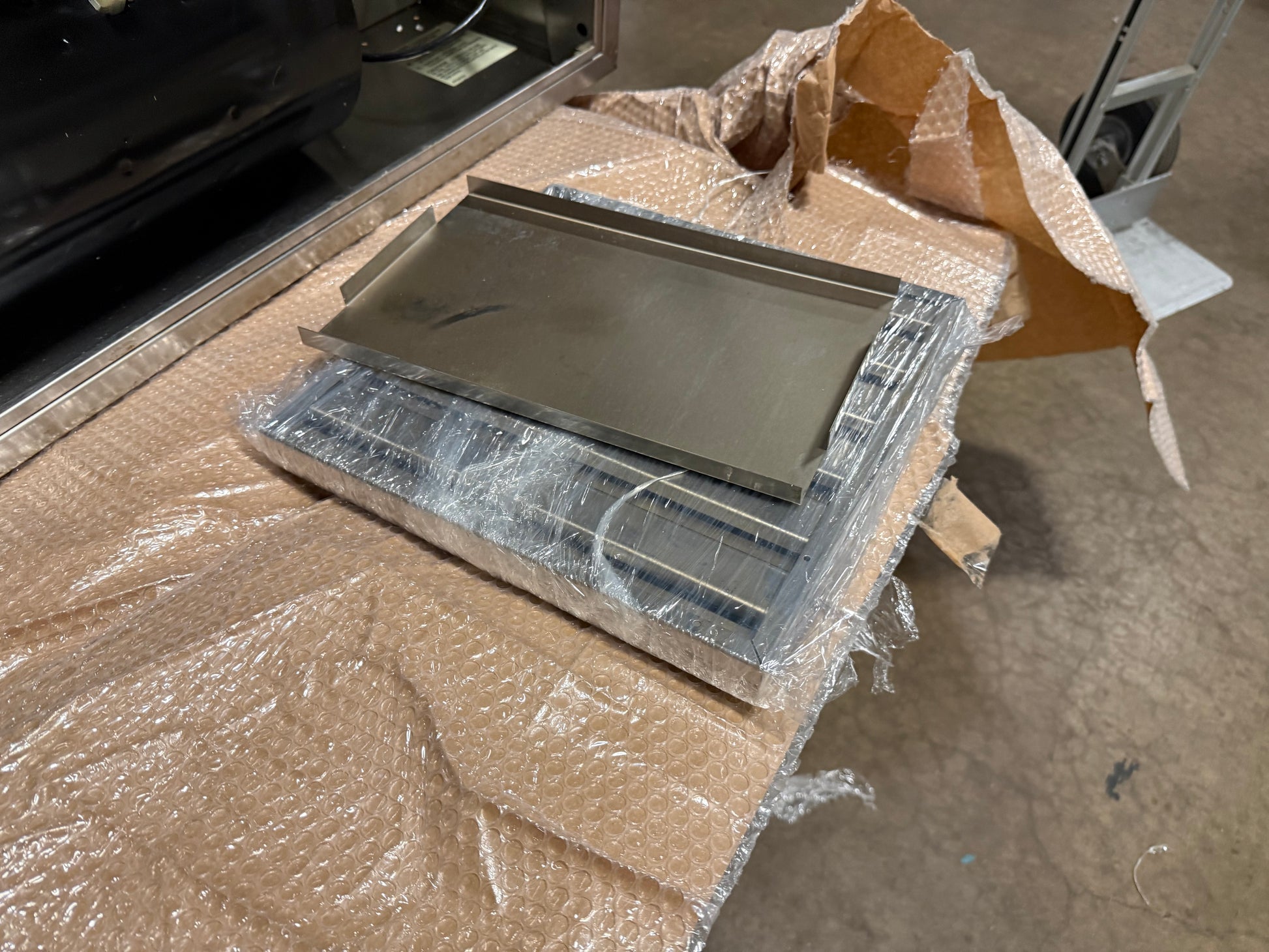 A stainless steel rectangular tray or griddle plate wrapped in clear plastic, resting on a cardboard box on a warehouse floor.