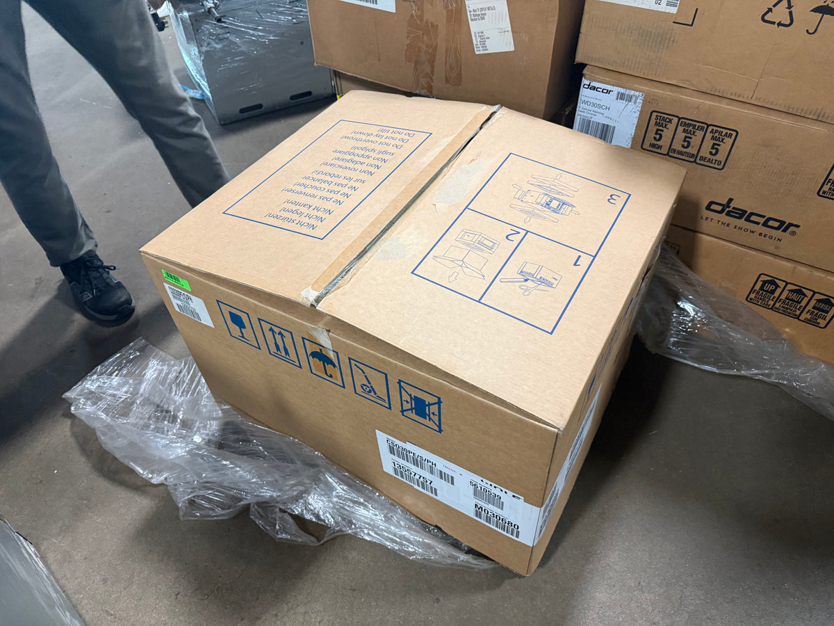 A large brown cardboard shipping box with blue printed handling instructions and a Dacor logo on a neighboring box, sitting on a concrete floor with plastic wrapping around it. A person's legs are partially visible in the upper left.