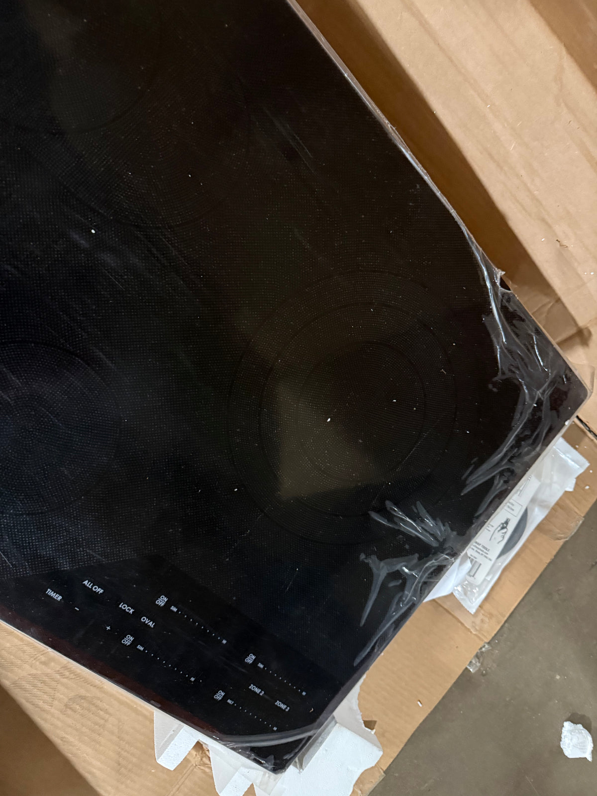 A black glass-ceramic induction cooktop with a control panel, still wrapped in protective plastic, resting on cardboard packaging.