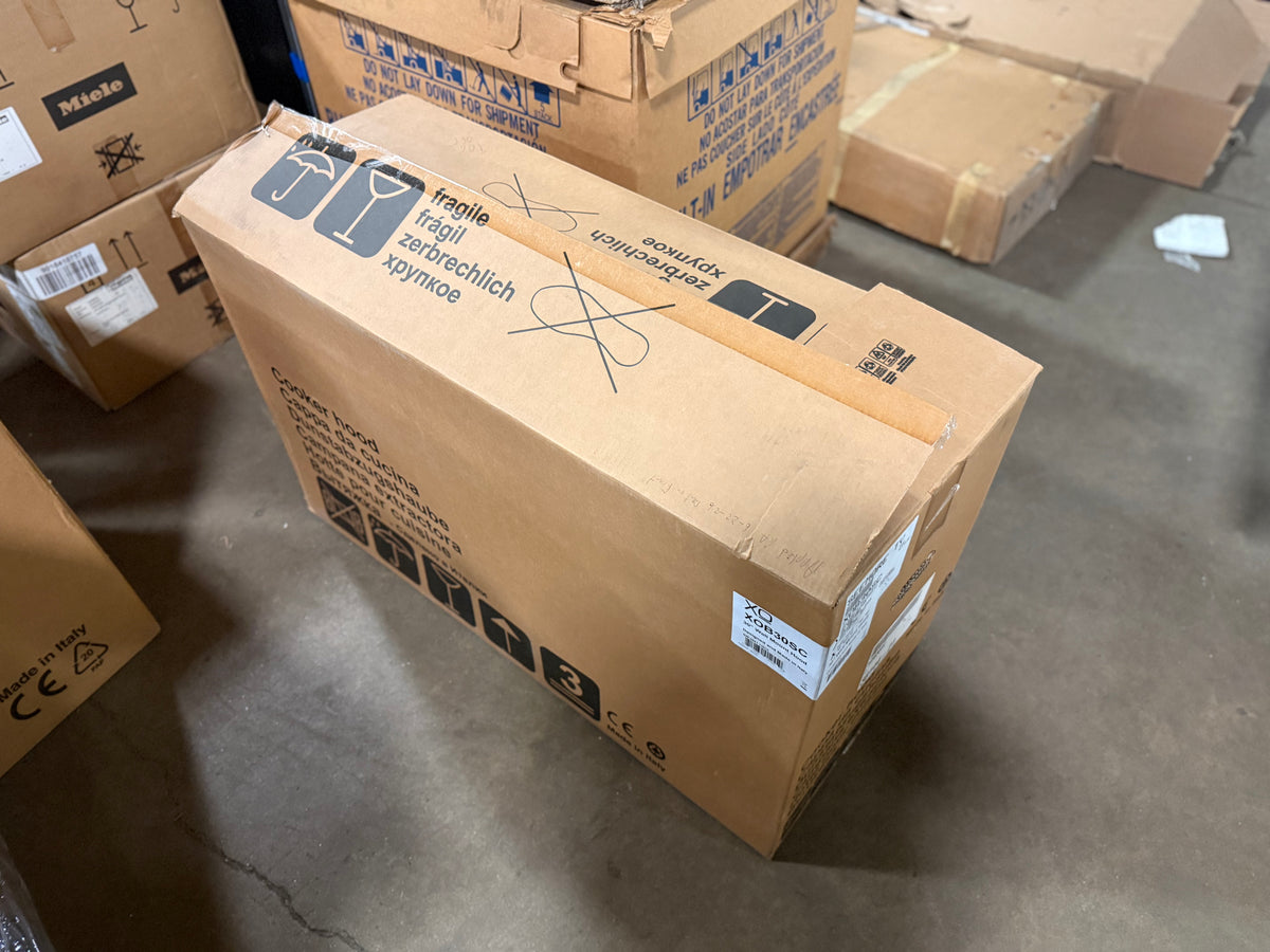 A brown cardboard shipping box with printed handling instructions and a white shipping label, photographed on a concrete floor among other stacked boxes. A Miele logo is visible on one of the surrounding boxes.