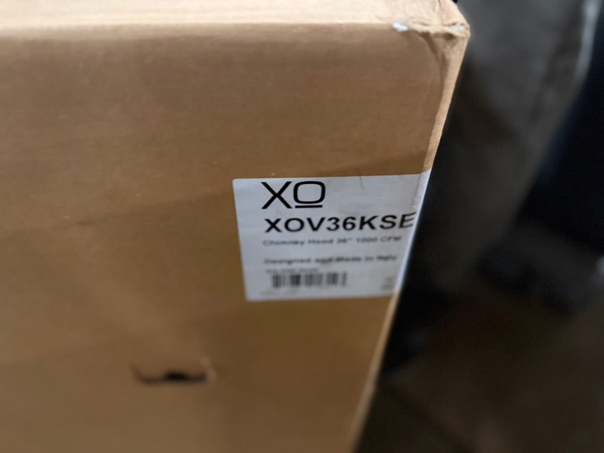 A close-up of a brown cardboard shipping box with a white label that reads XO XOV36KSE, photographed in a real environment with a blurred background.