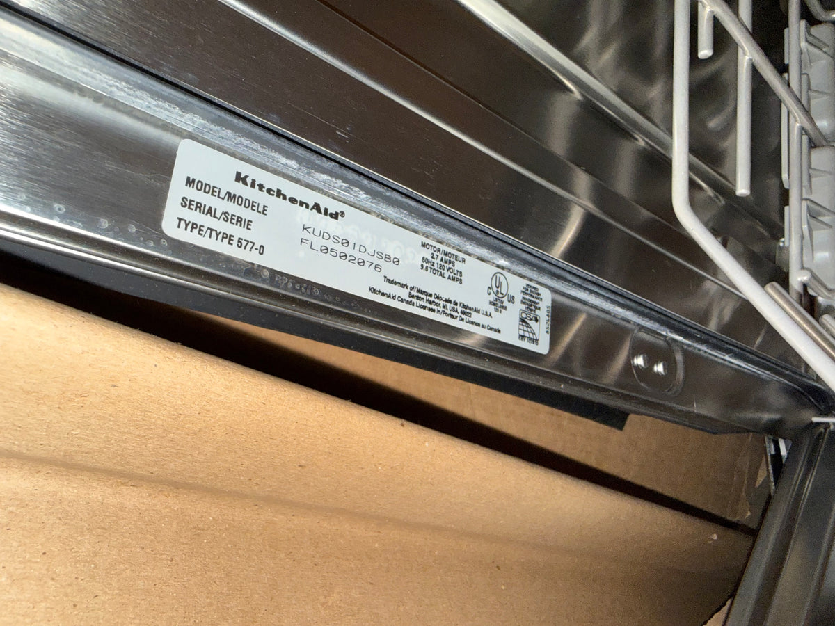 A close-up of a KitchenAid dishwasher interior showing the model label on the metal trim, with cardboard packaging visible below.