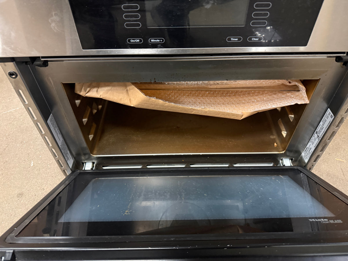 An open stainless steel built-in oven with its door lowered, showing a brown paper bag or liner inside on the rack. The oven door has a black glass panel and a control interface is partially visible at the top.