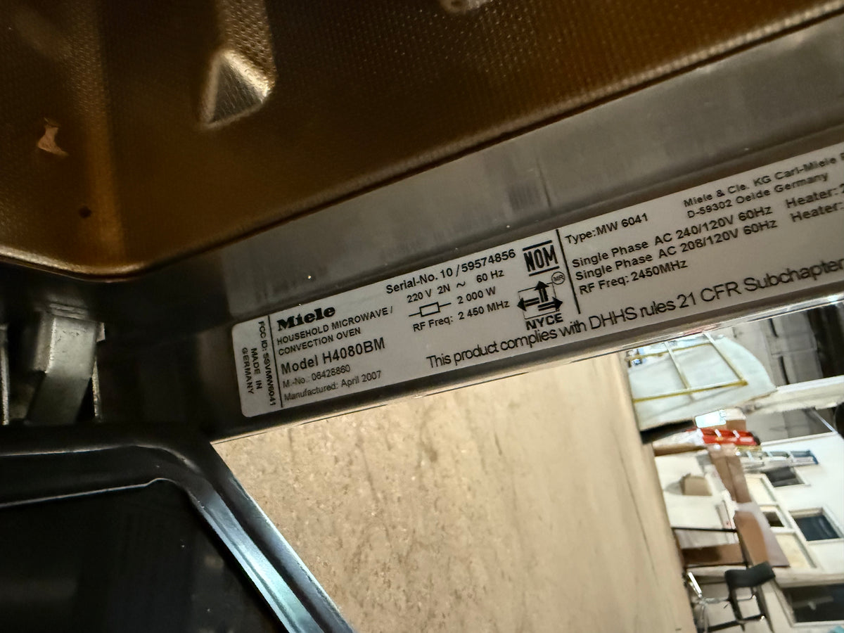 A close-up of a product label on a metallic appliance trim, showing the Miele logo and model information H4080BM, with a workshop or showroom environment visible in the background.