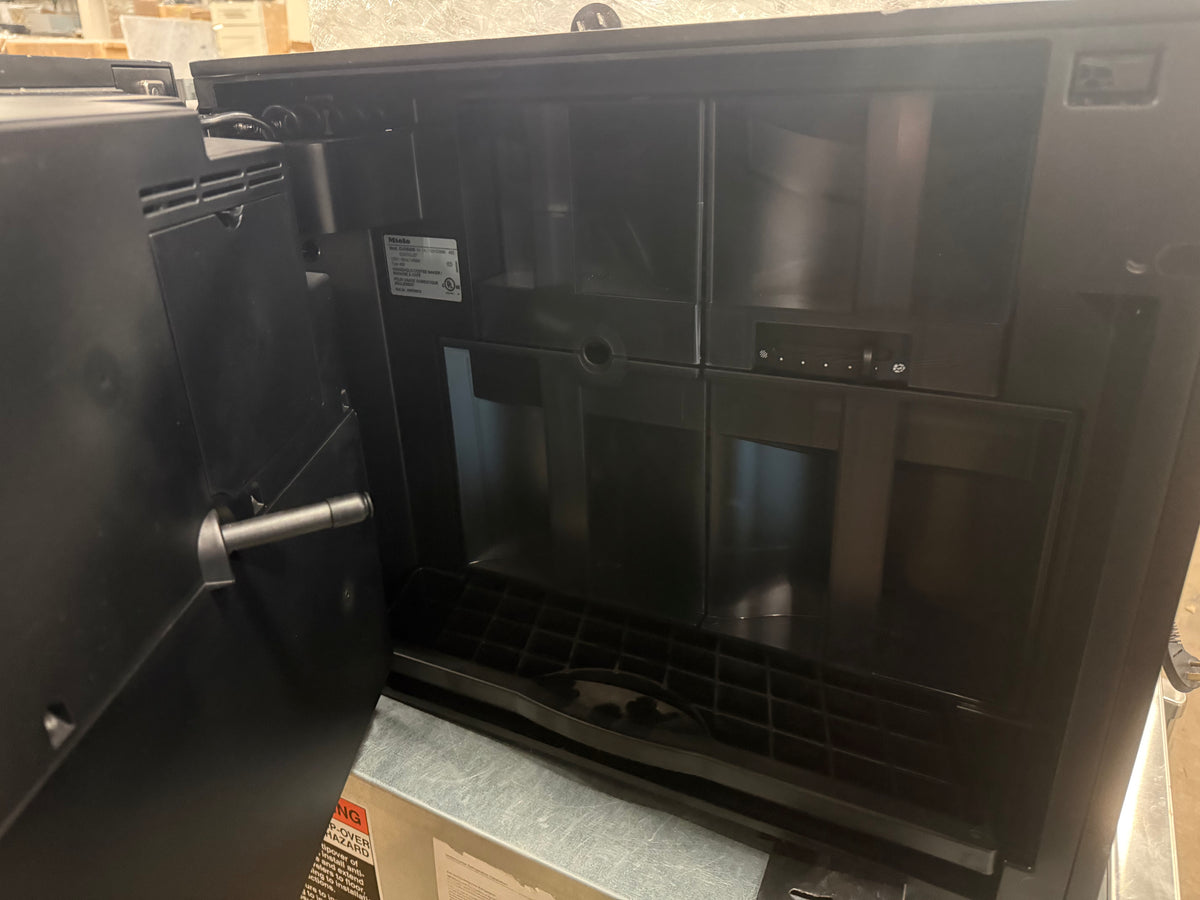 A black countertop coffee maker with its door open, showing the interior drip tray and upper chamber, photographed in what appears to be a warehouse or showroom setting.