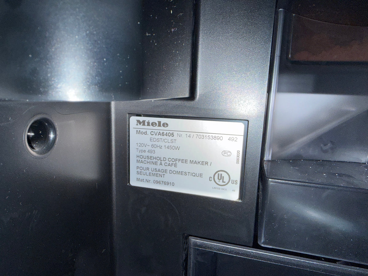 Close-up of a Miele household coffee maker label on the side panel of a dark appliance, showing model CVA8406 and power details.
