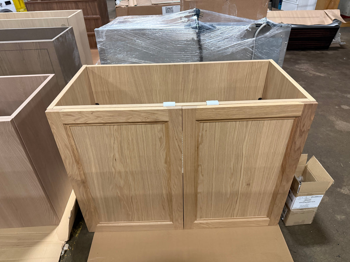 A light oak wood double cabinet base with paneled doors, shown in a warehouse setting with other furniture pieces and cardboard boxes around it.