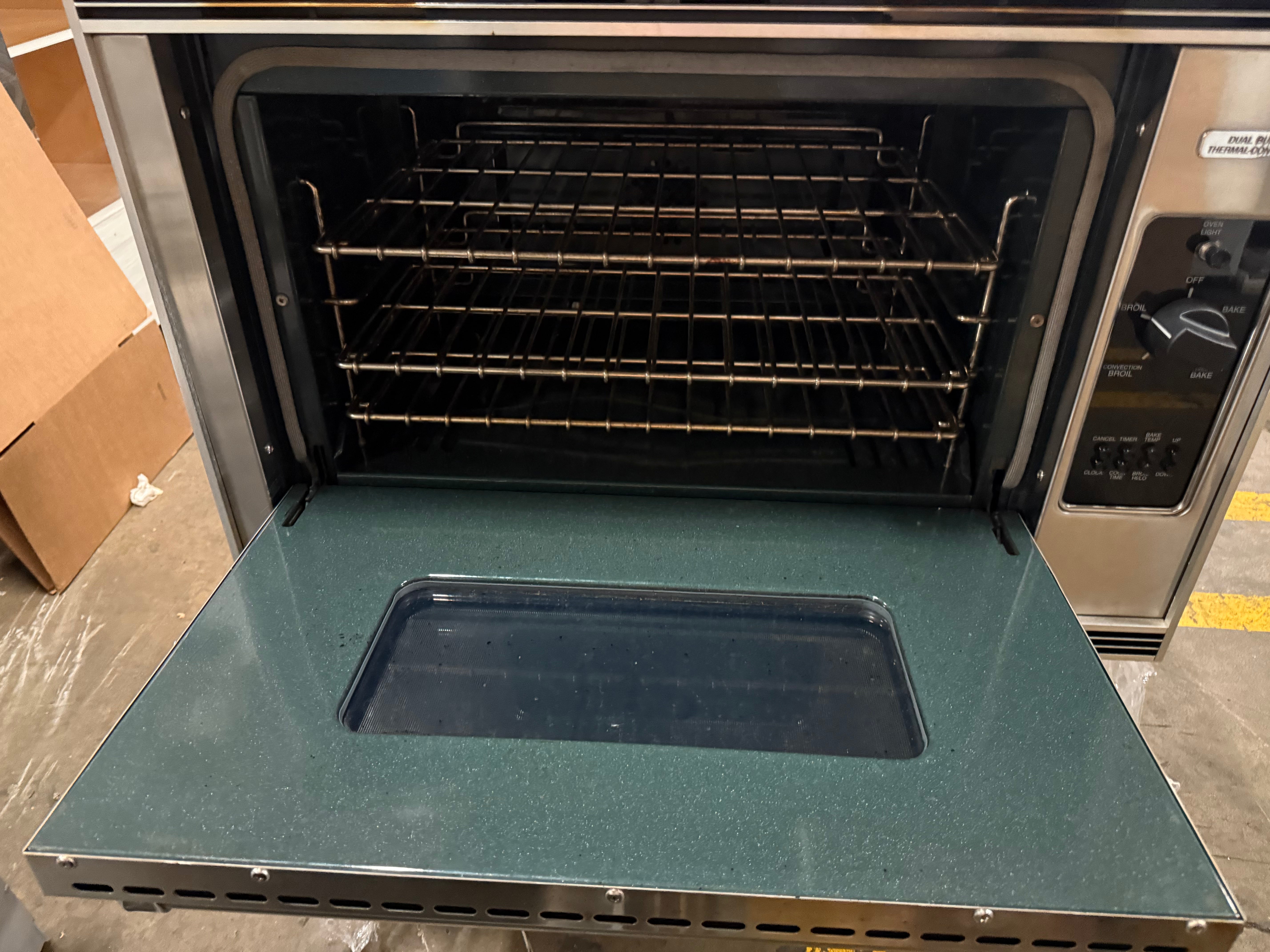 A stainless steel countertop oven with its door open, showing three interior racks and a dark green speckled door panel with a recessed tray insert.