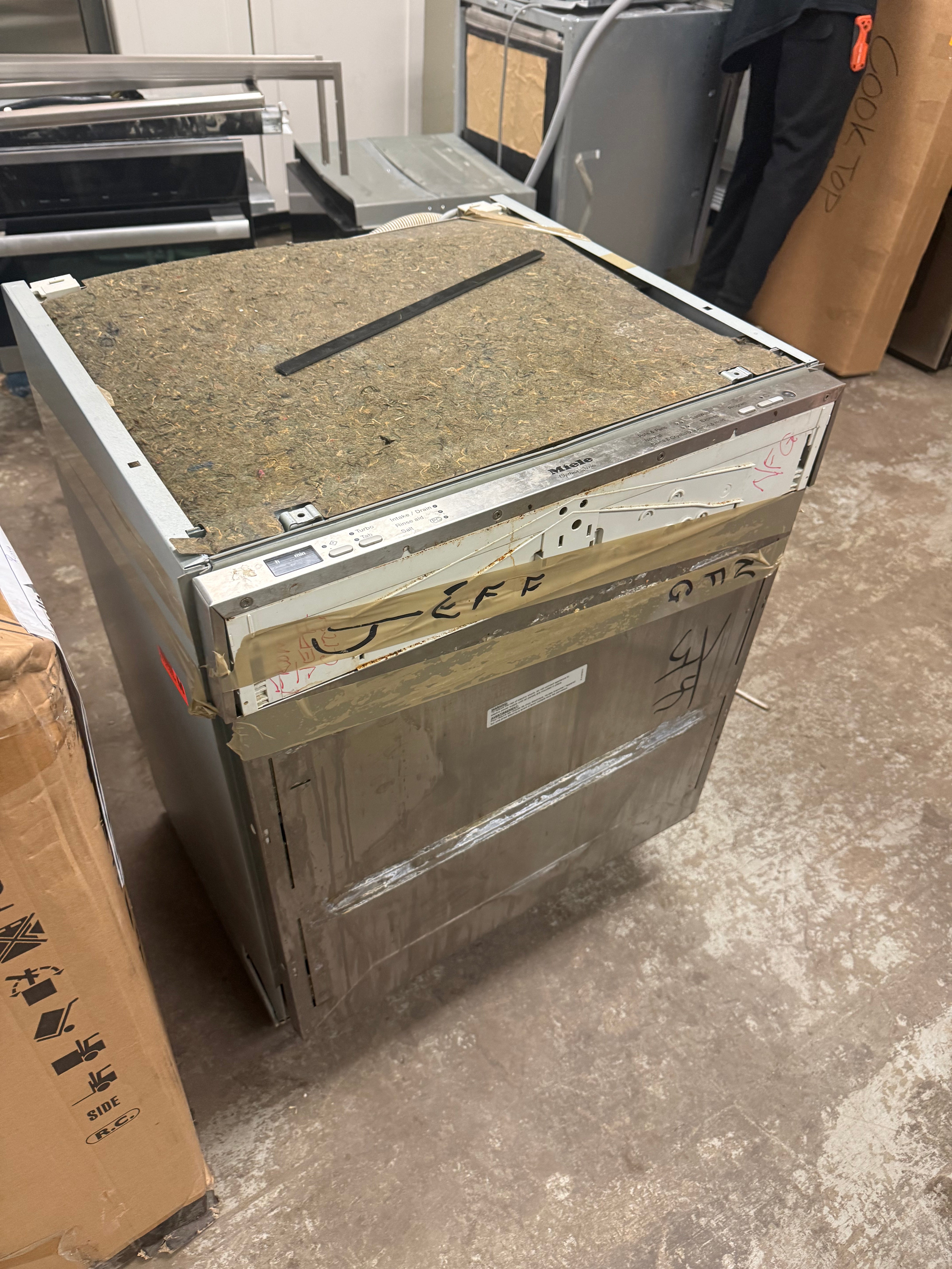 A large appliance, likely a dishwasher or similar countertop machine, wrapped in protective cardboard and tape in a warehouse setting. The top surface is covered with a brown textured mat, and handwritten markings are visible on the front.