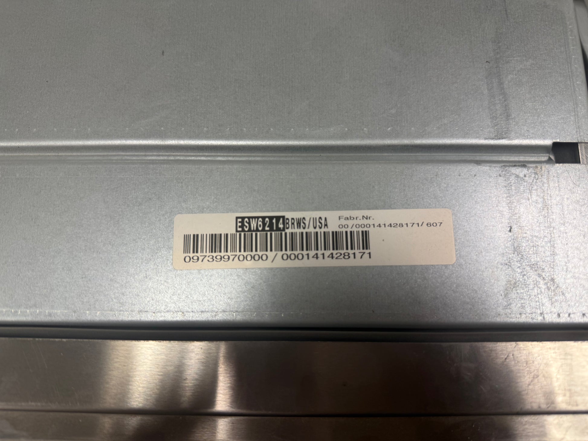 Close-up of the underside of a metallic silver appliance or device, showing a white barcode label with printed text and numbers.