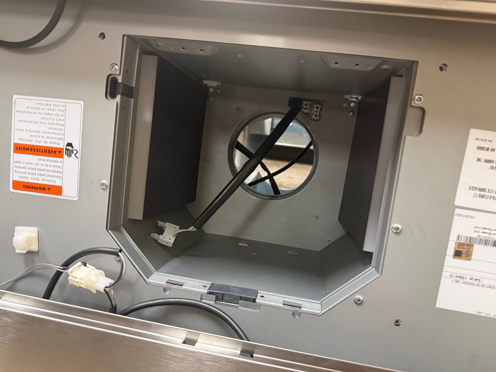 Close-up of the interior vent opening of a stainless steel appliance, showing a black fan blade, wiring, and warning labels on a gray metal panel.