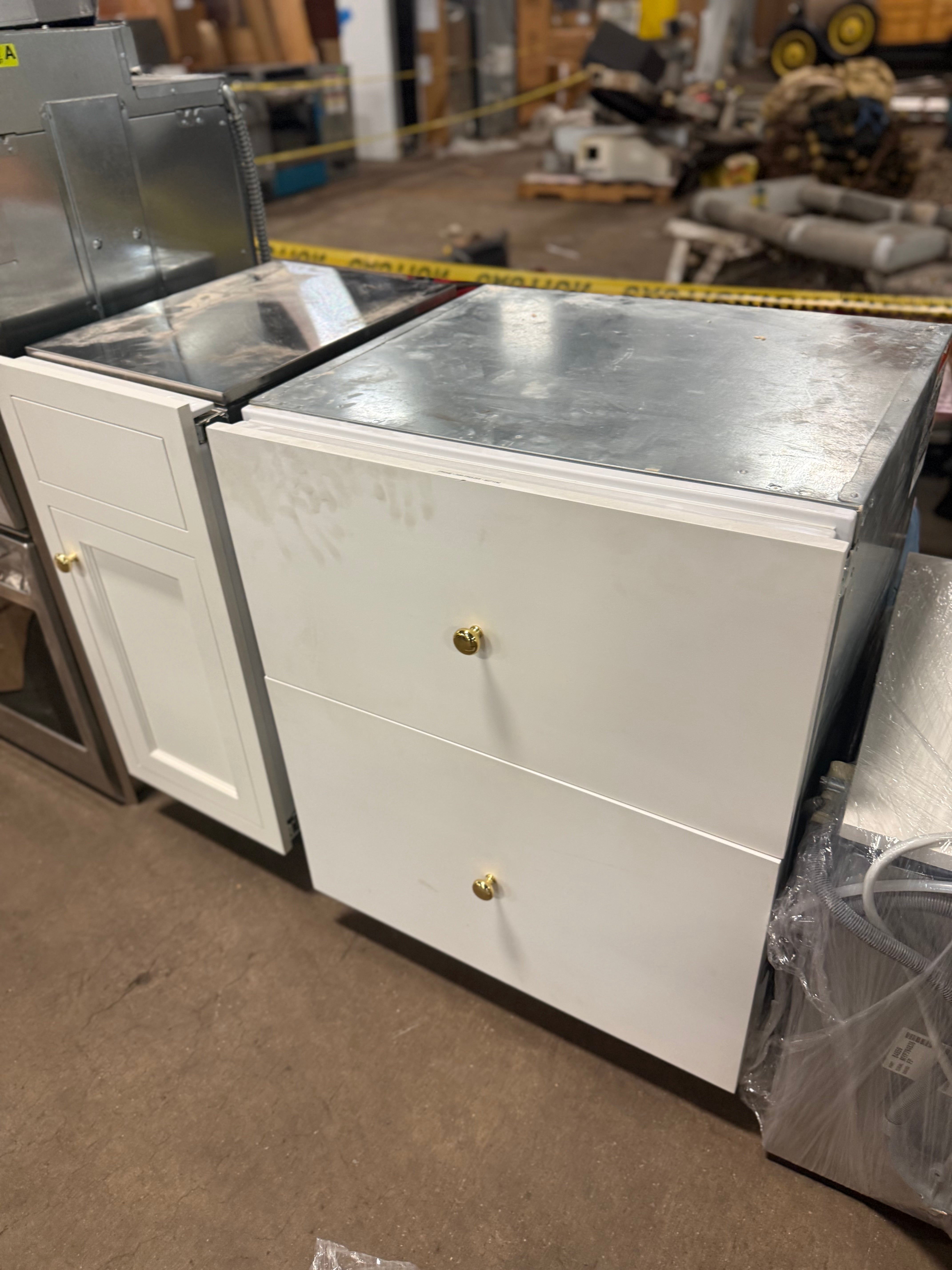 A white two-drawer kitchen cabinet with a brushed stainless steel top and small round gold knobs, photographed in a warehouse or showroom setting.