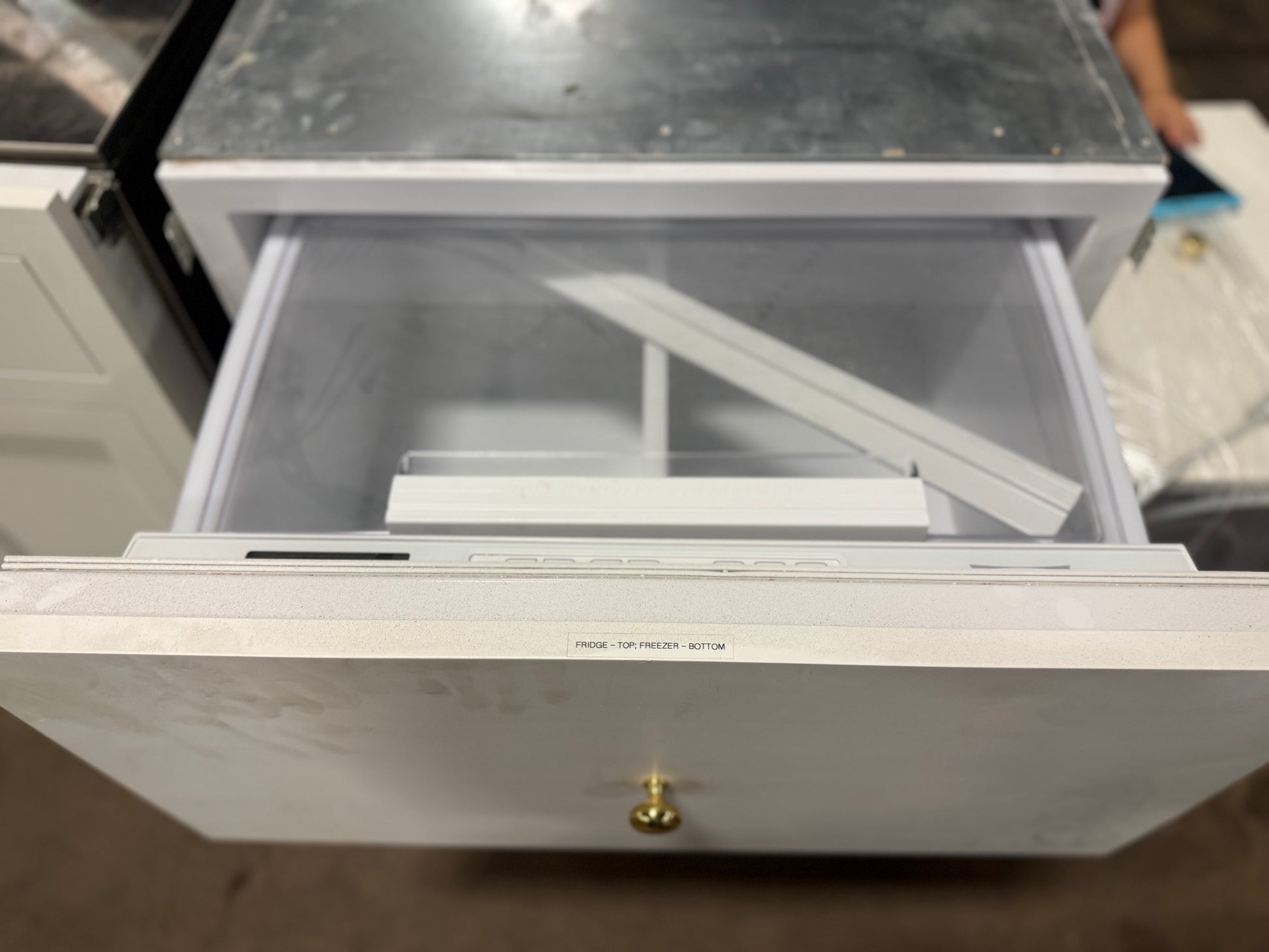 A white drawer with a small brass knob, shown partially open to reveal a gray interior organizer tray. The top surface of the unit is dark gray, and the background is a cluttered workshop or warehouse floor with other items visible.