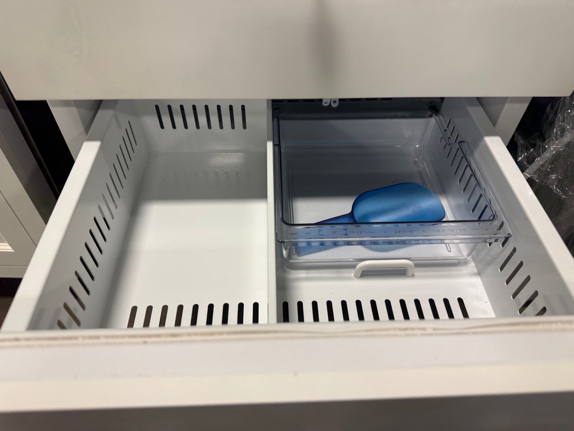 An open refrigerator drawer with two interior bins: a larger empty white bin on the left and a smaller clear bin on the right containing a blue scoop, set inside a white refrigerator.
