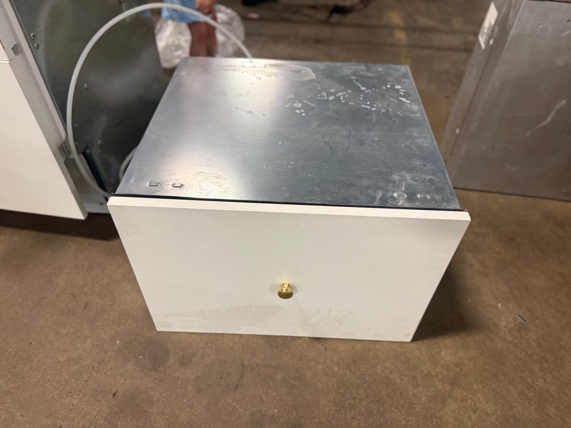 A white metal utility drawer with a brushed silver metal top lid and a small brass lock, shown on a concrete floor.