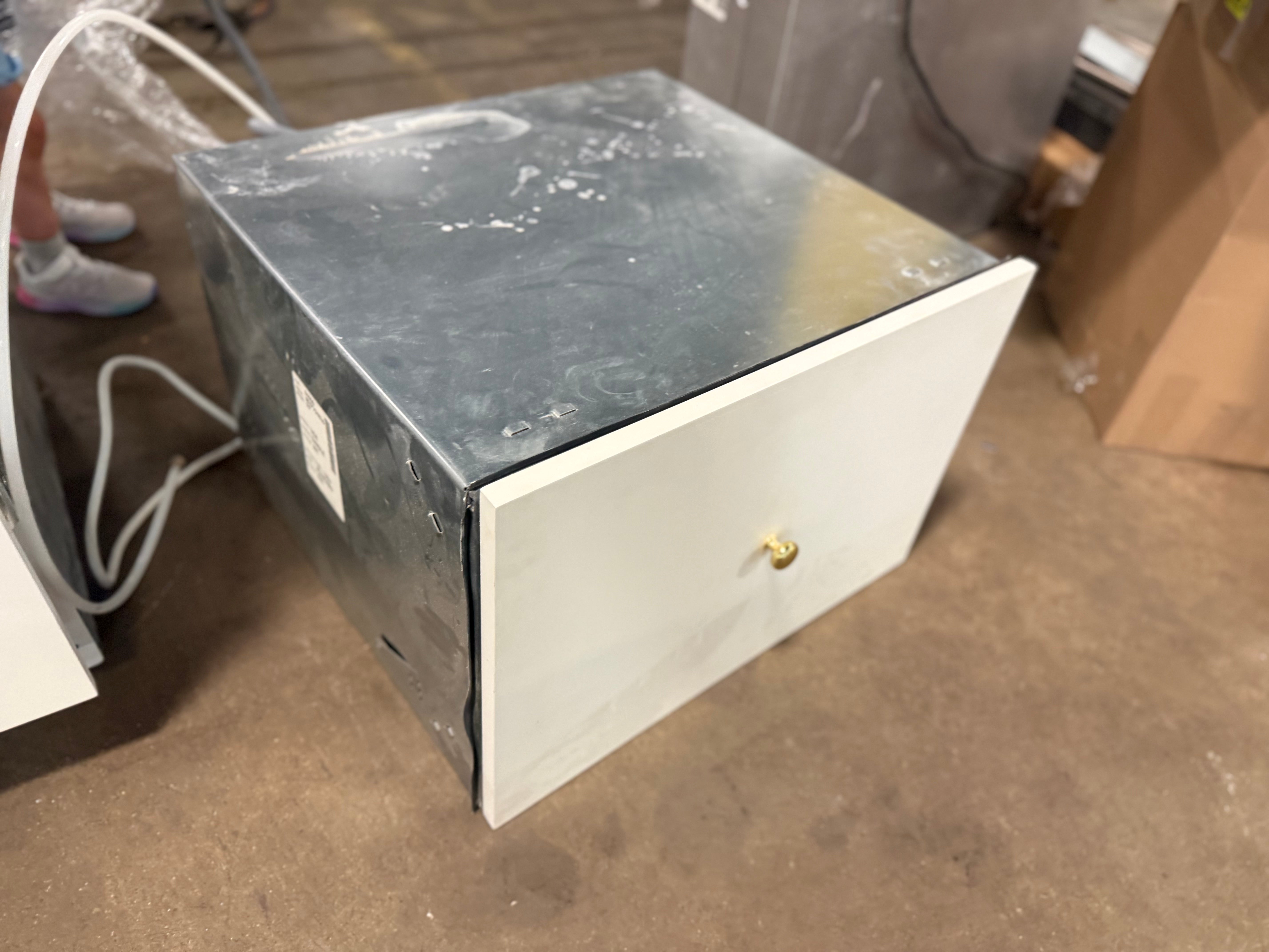A white and dark gray metal storage cabinet or drawer with a small brass knob, sitting on a concrete floor in what appears to be a warehouse or workshop.