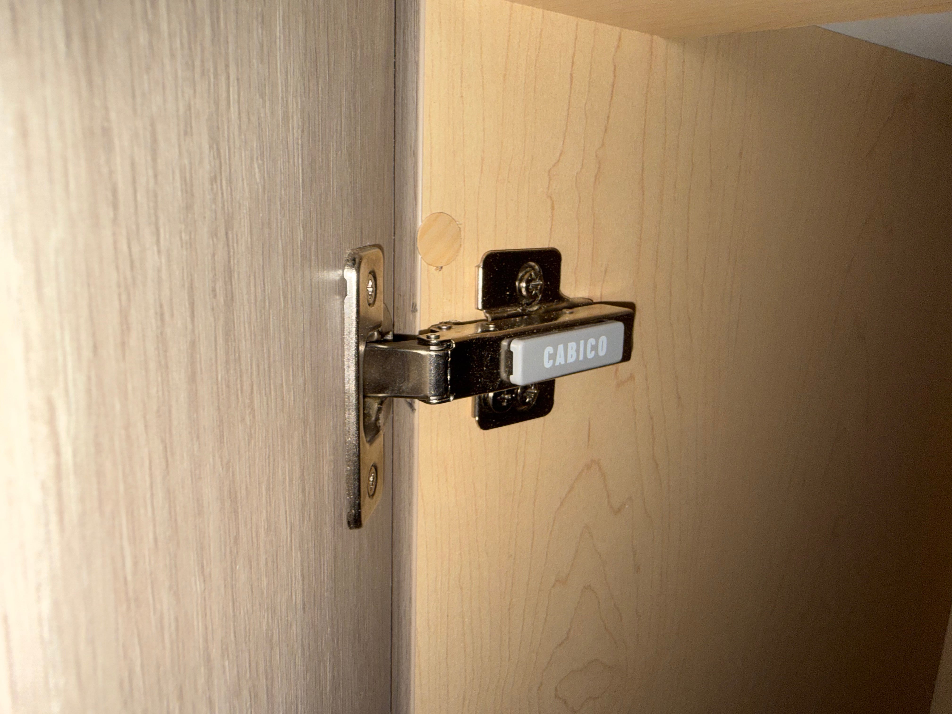 A close-up of a hinge installed on a light wood cabinet door, with a white label on the hinge that reads CABICO.
