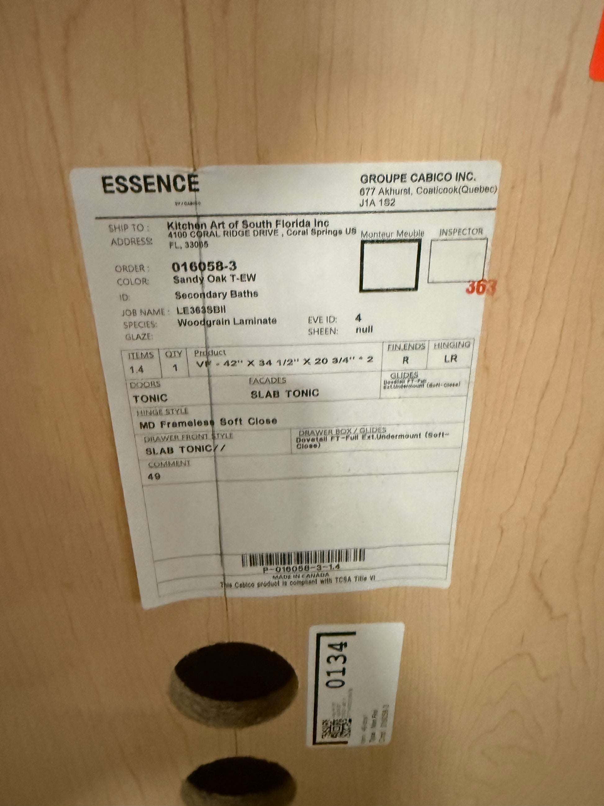 A white paper product label for ESSENCE by Groupe Cabrio Inc. is taped onto a light wood surface, with a black circular object partially visible below it.