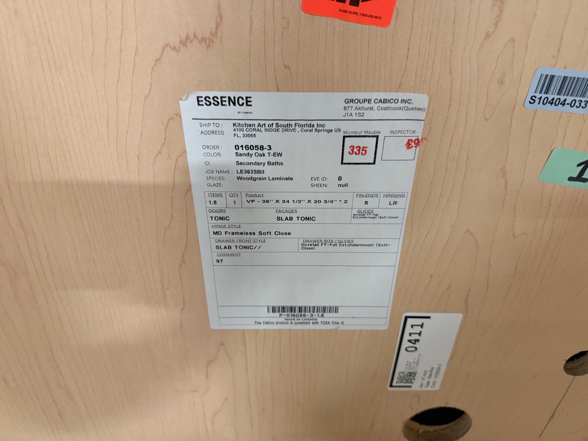 A white product information label for an ESSENCE bathroom vanity cabinet, showing order details, a red price of $335, and a slat count of 3, attached to a light wood cabinet door.