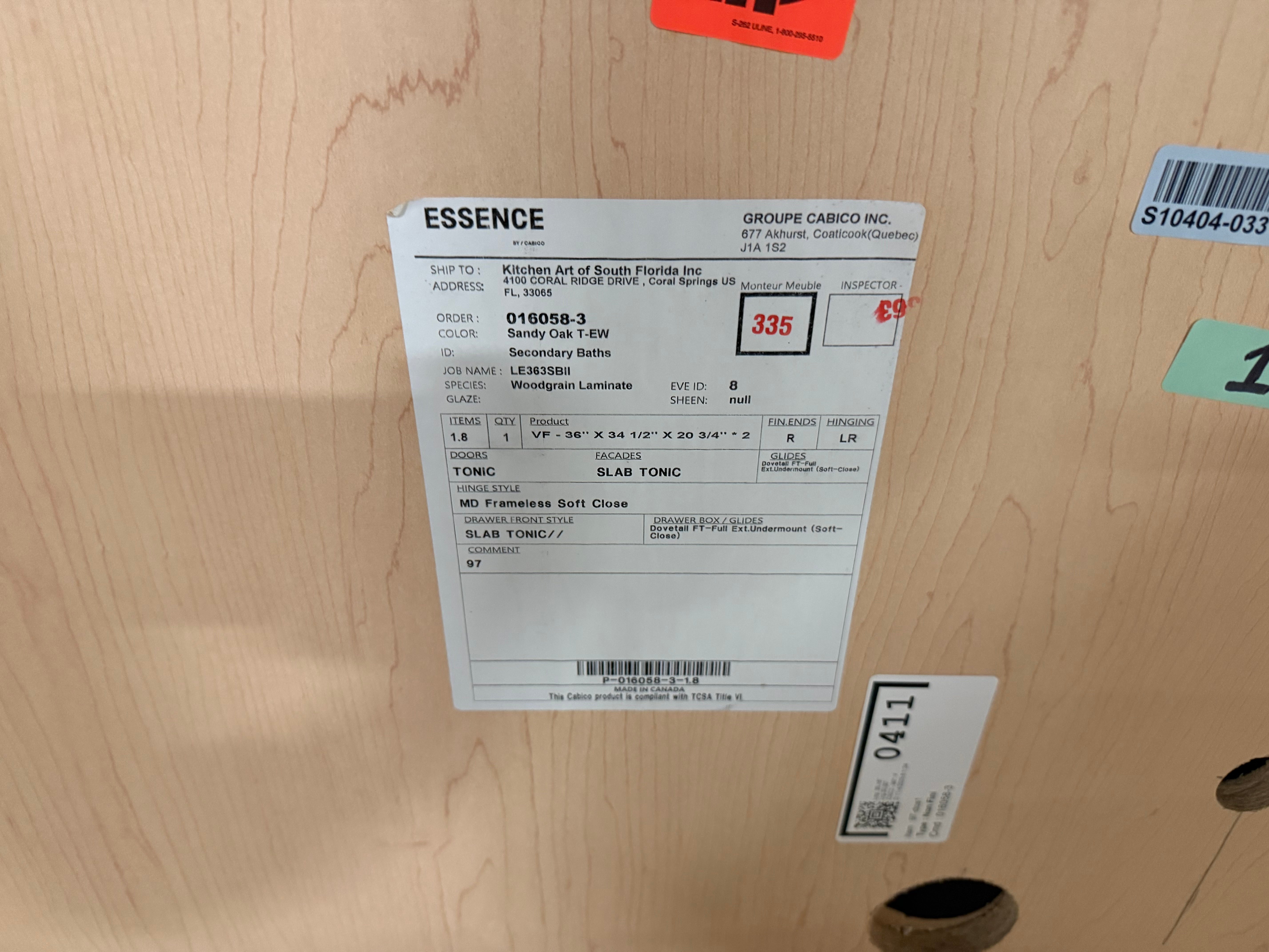 A white product information label for an ESSENCE bathroom vanity cabinet, showing order details, a red price of $335, and a slat count of 3, attached to a light wood cabinet door.
