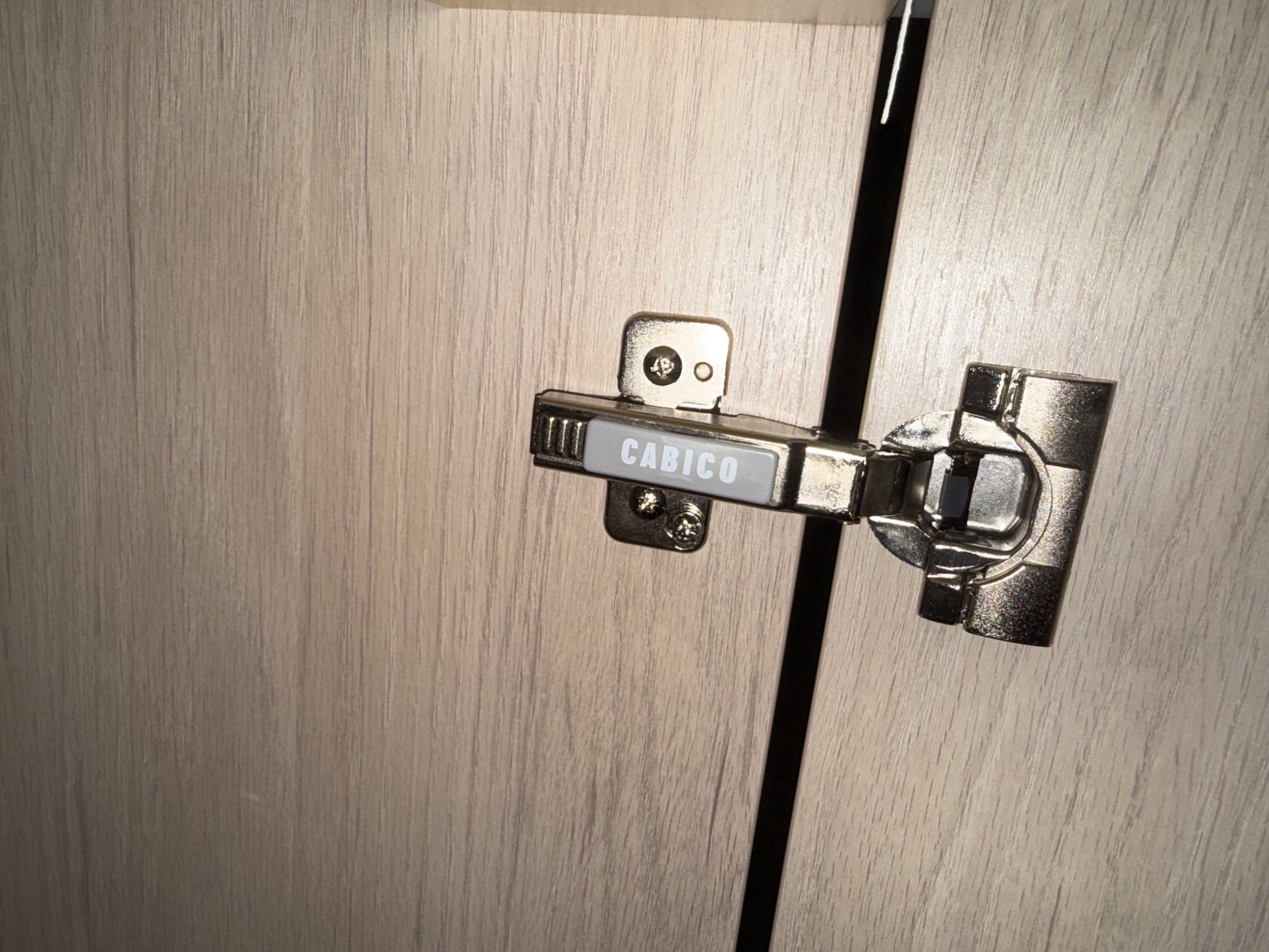 A close-up of a silver metal cabinet hinge with the brand name CABICO visible on its center label, mounted on light wood grain cabinet doors.