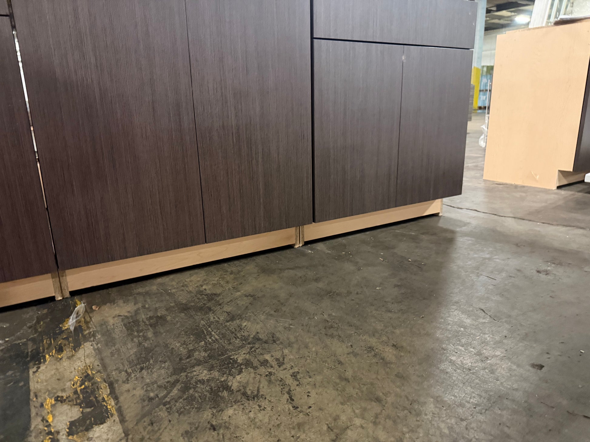 Dark brown wood grain cabinet doors with light wood bases, shown in a warehouse setting on a concrete floor.