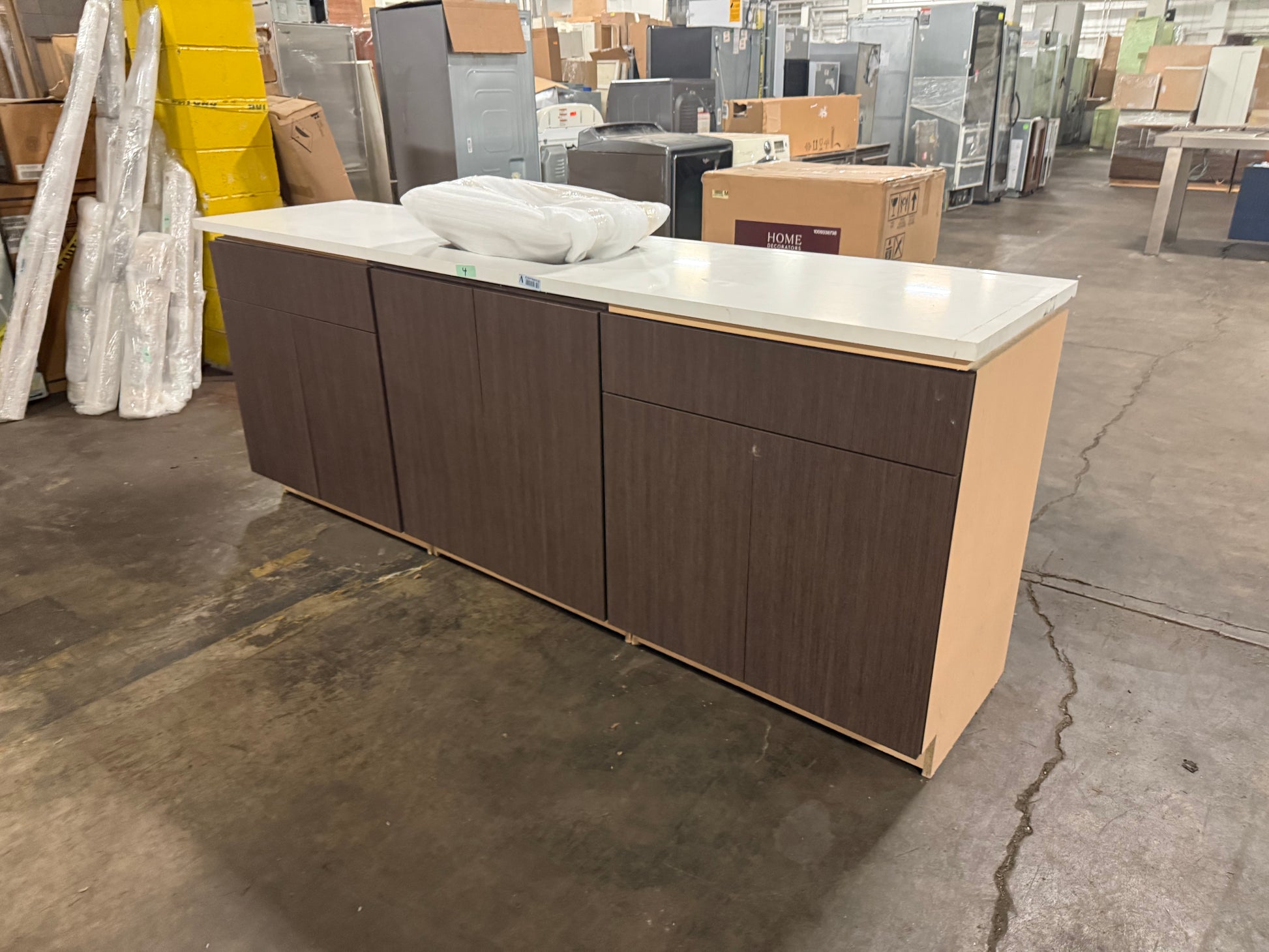 A long kitchen or bar cabinet unit with dark brown wood-look doors and a white countertop, photographed in a warehouse or furniture store setting with other appliances and boxes in the background.