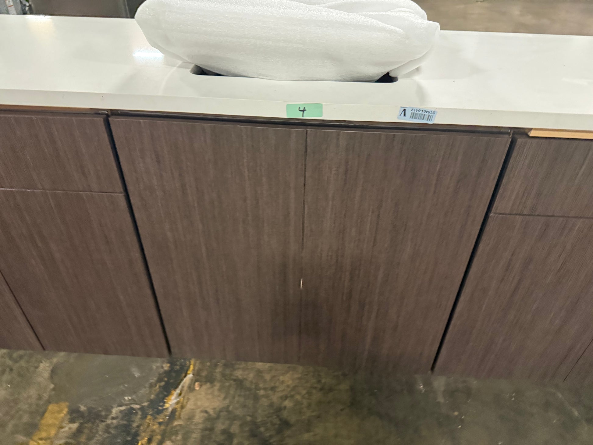 A close-up of a modern floating vanity cabinet with dark brown wood grain doors and a white countertop. A white towel is draped over the top edge, and small inventory stickers are visible on the front.