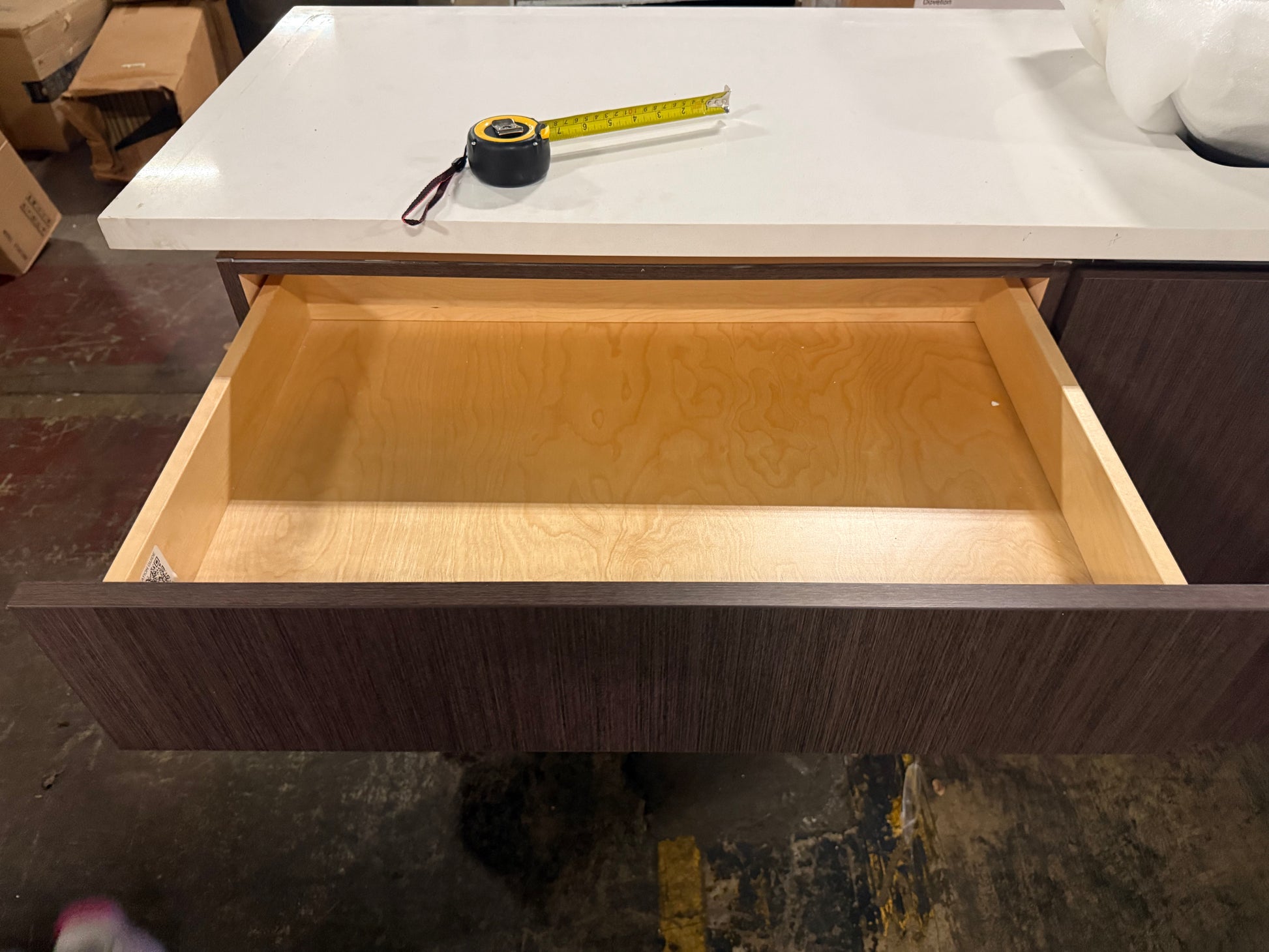 A dark wood grain drawer with an open light wood interior, shown below a white countertop with a tape measure resting on it.